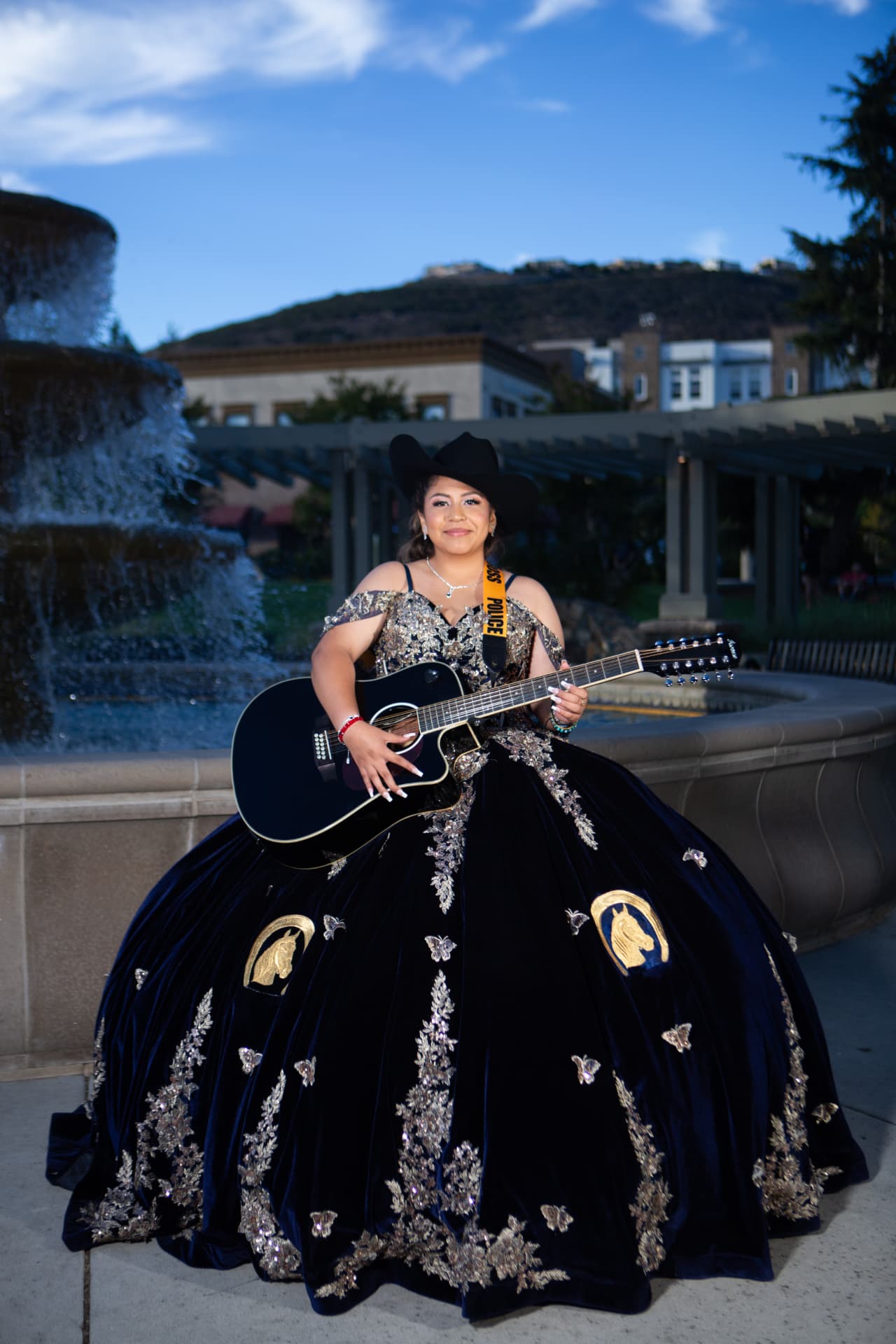 Quinceañera in a navy velvet gown and black hat holding a twelve-string guitar by a fountain