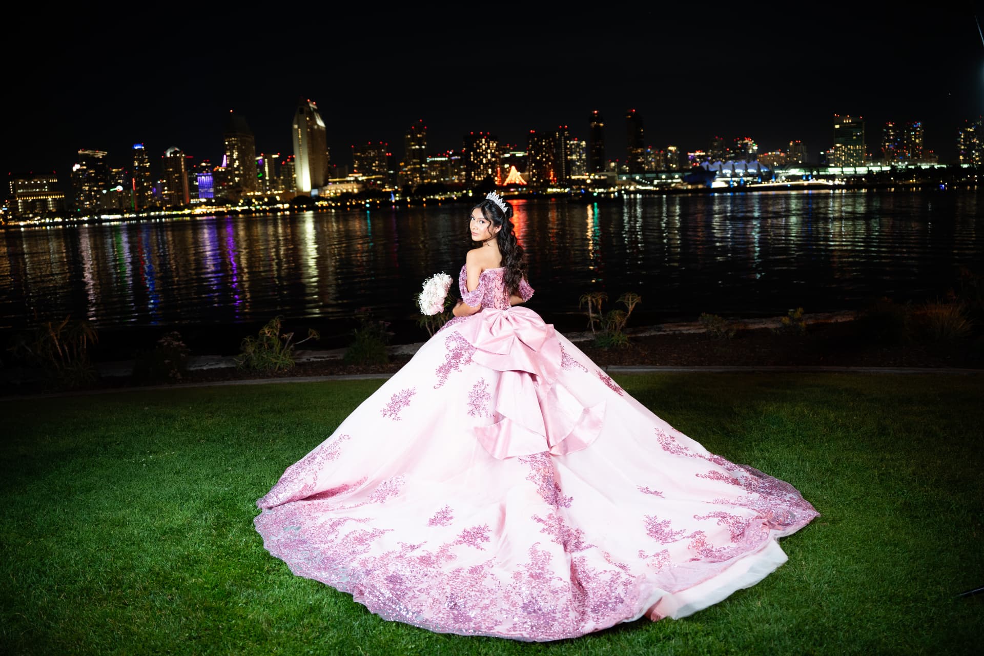 Quinceañera in a pink gown seen from behind with her bouquet against the glittering San Diego skyline at night