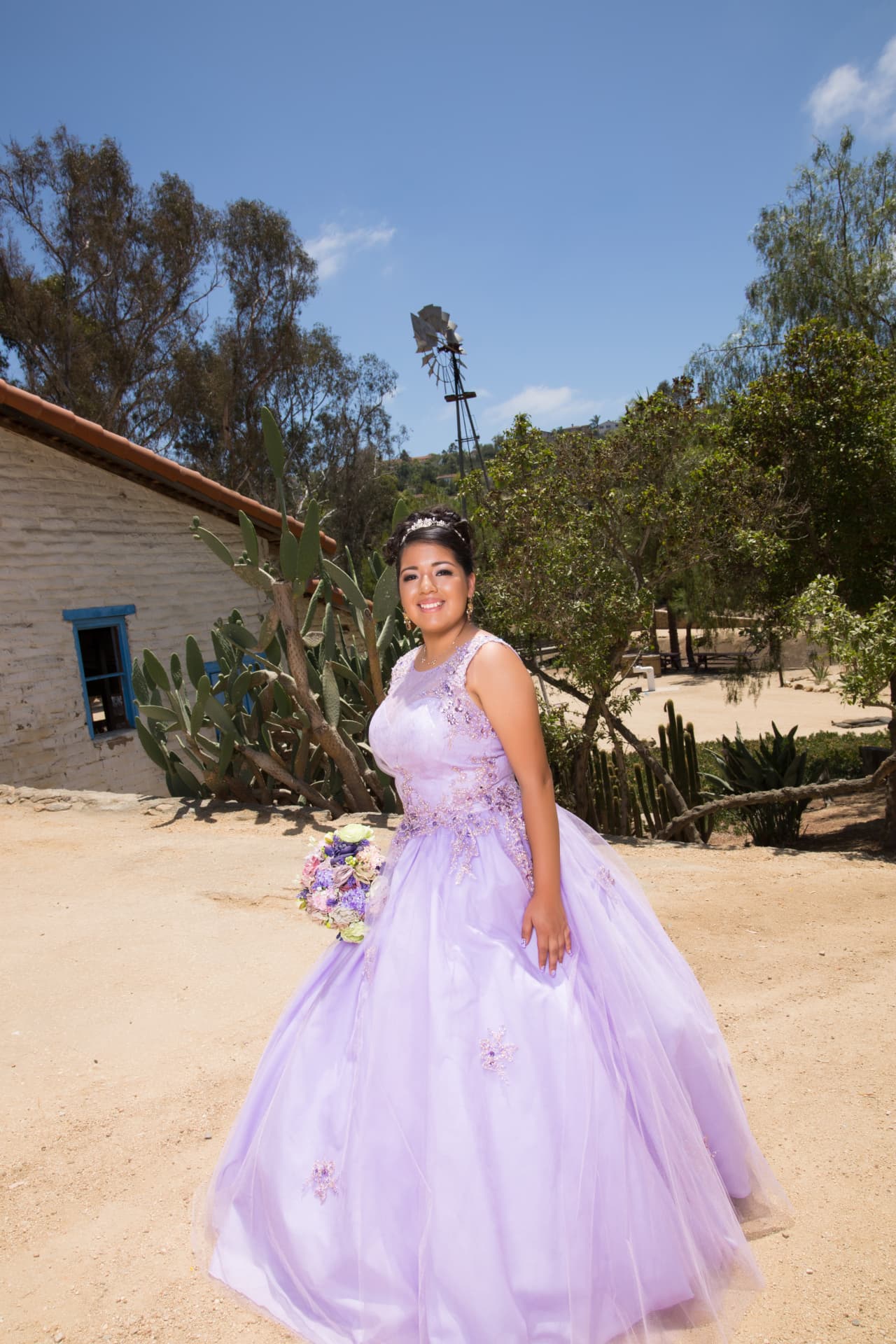 Quinceañera in a lavender gown holding a bouquet at a rustic ranch with cacti and a windmill