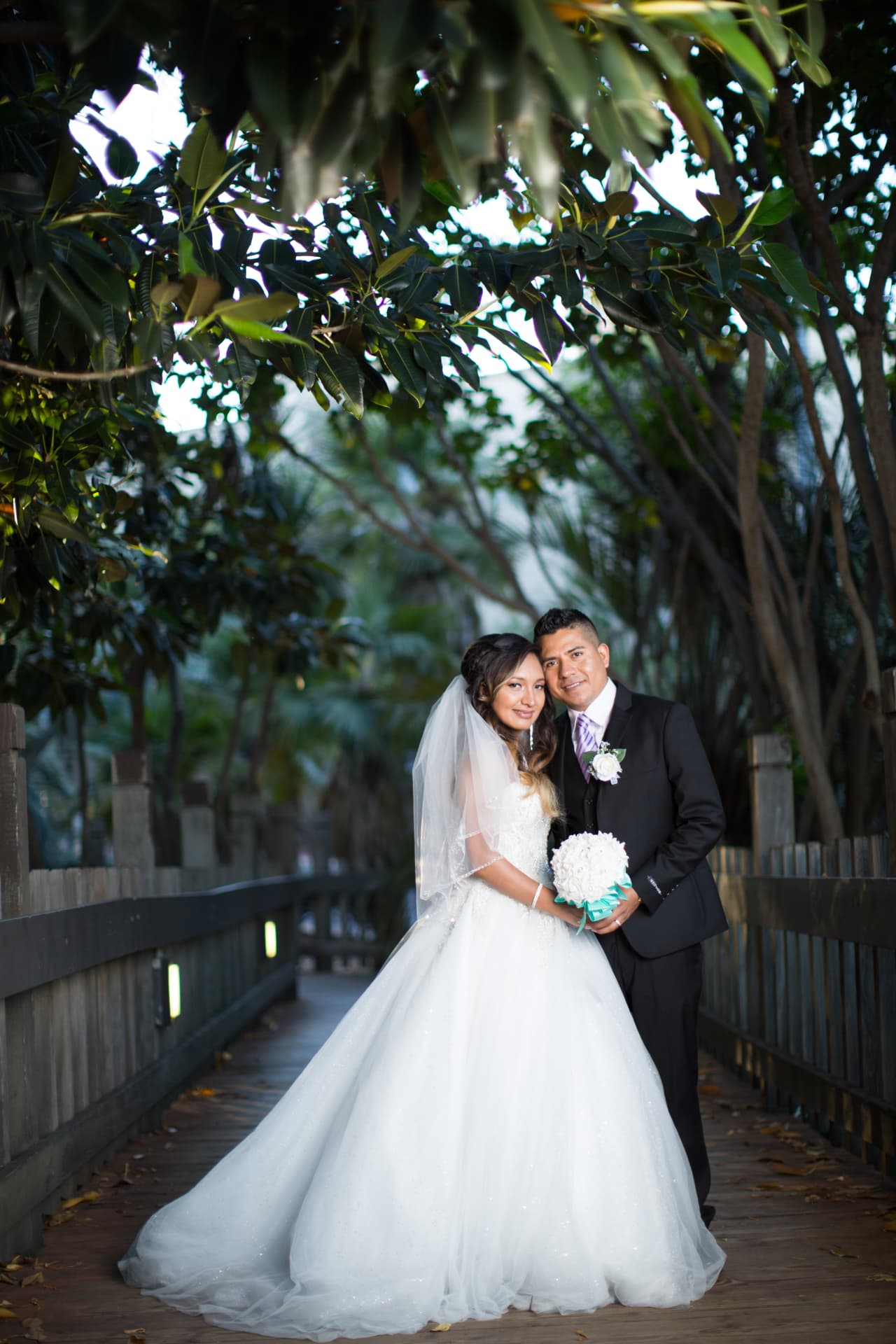 Bride and groom standing close together beneath leafy trees on a wooden boardwalk