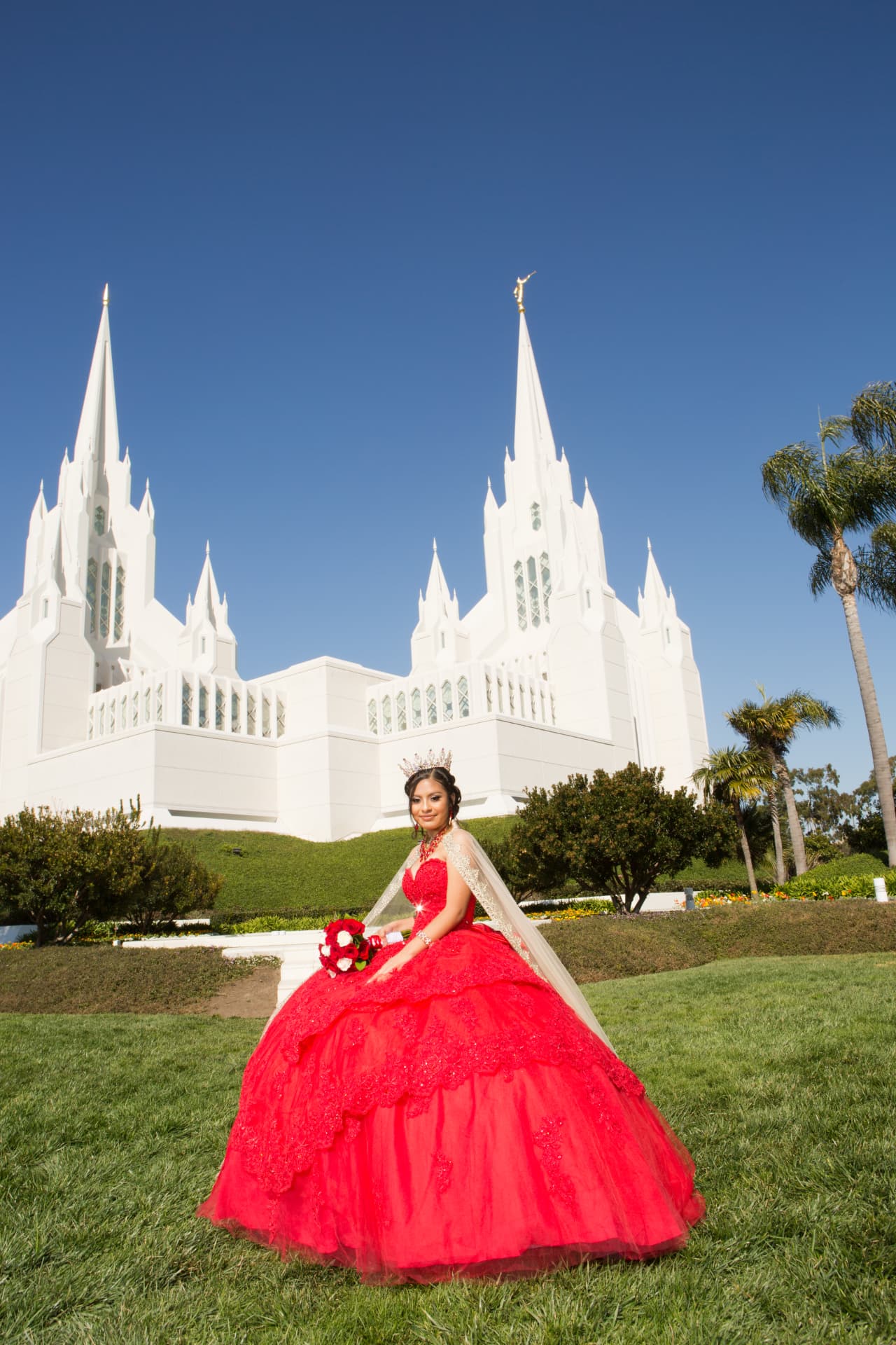Quinceañera in a red gown on a lawn in front of white temple spires beneath a clear blue sky