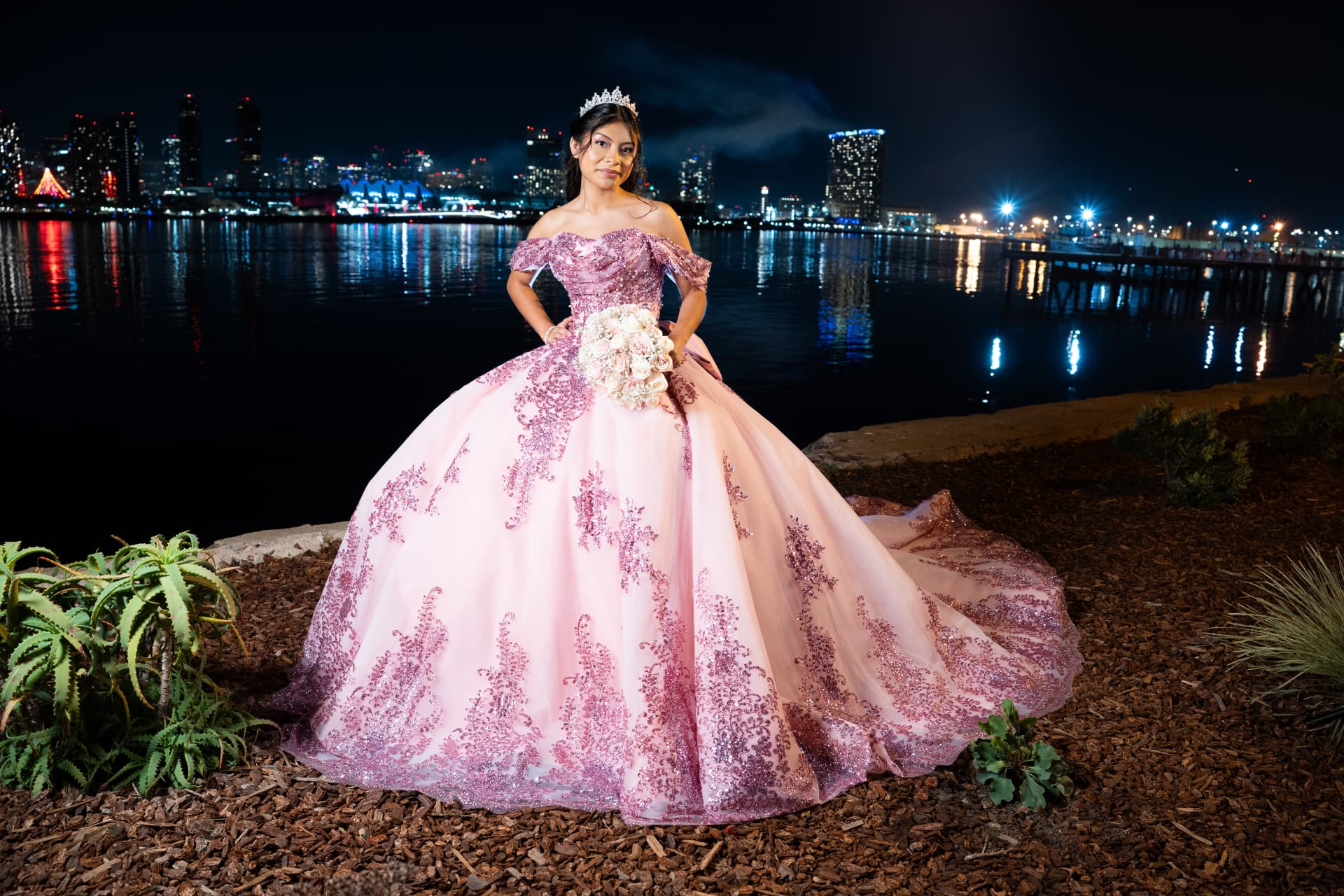 Quinceañera in a sparkling pink gown with her bouquet before the waterfront skyline at night