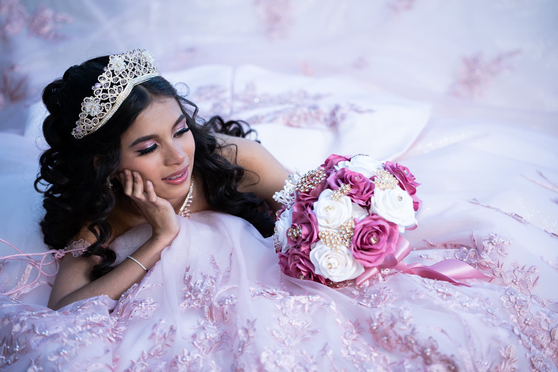 Quinceañera in a pink gown lying among her tulle skirt with her jeweled bouquet