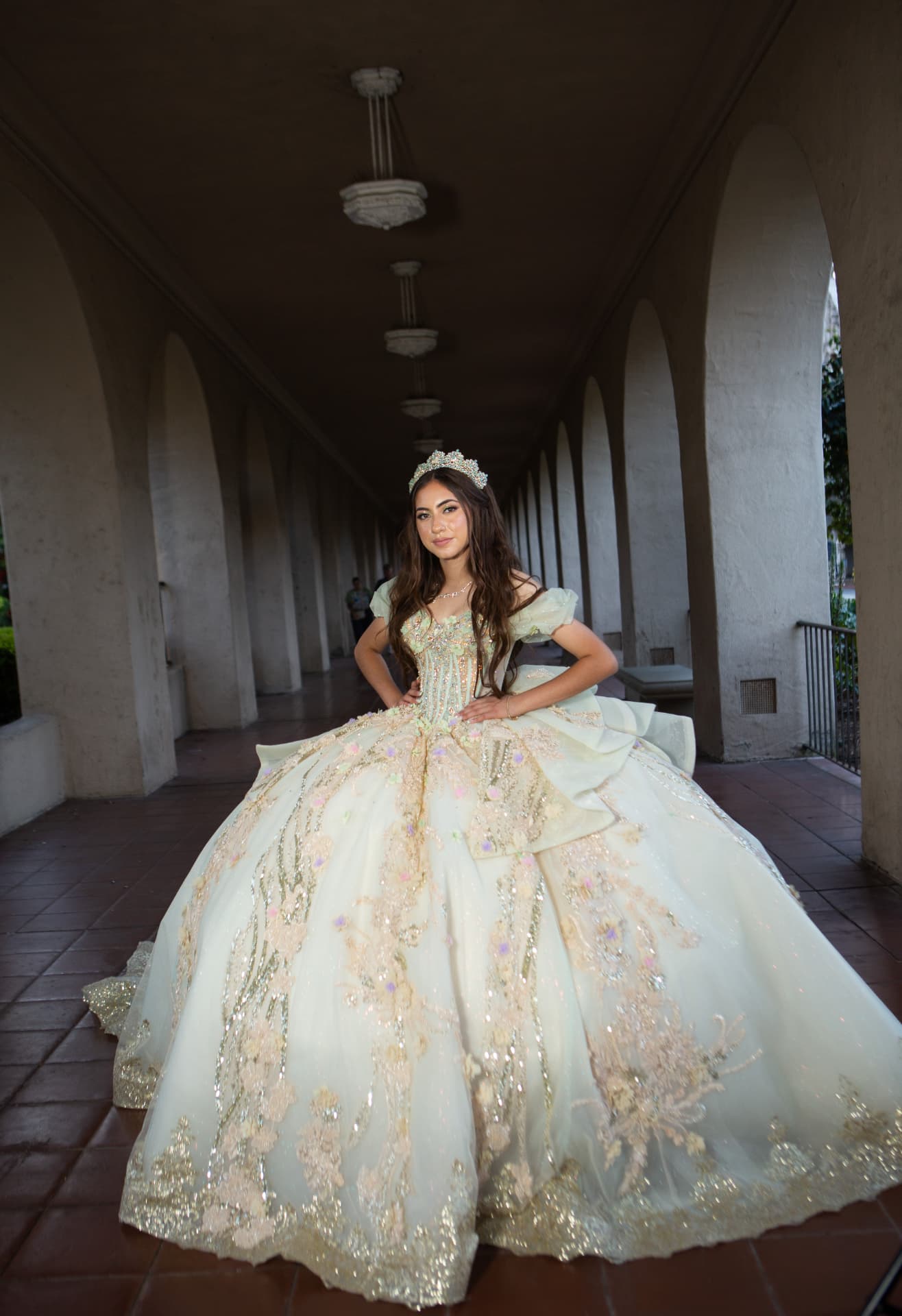 Quinceañera in a cream and gold gown posed hands-on-hips beneath a colonnade of arches