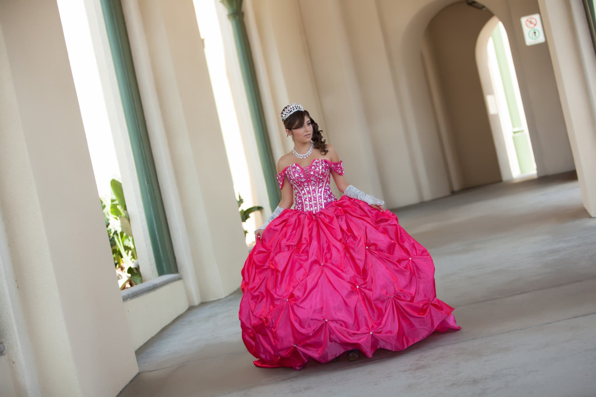 Quinceañera in a pink jeweled gown holding out her skirt in an arched corridor