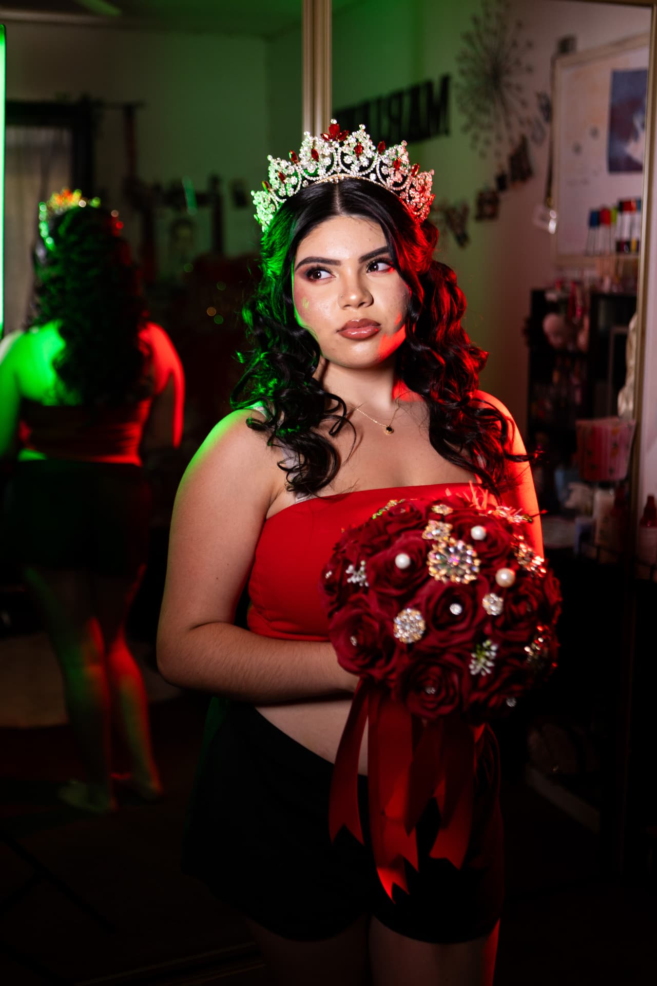 Quinceañera with her tiara holding a jeweled red rose bouquet under red and green party lights
