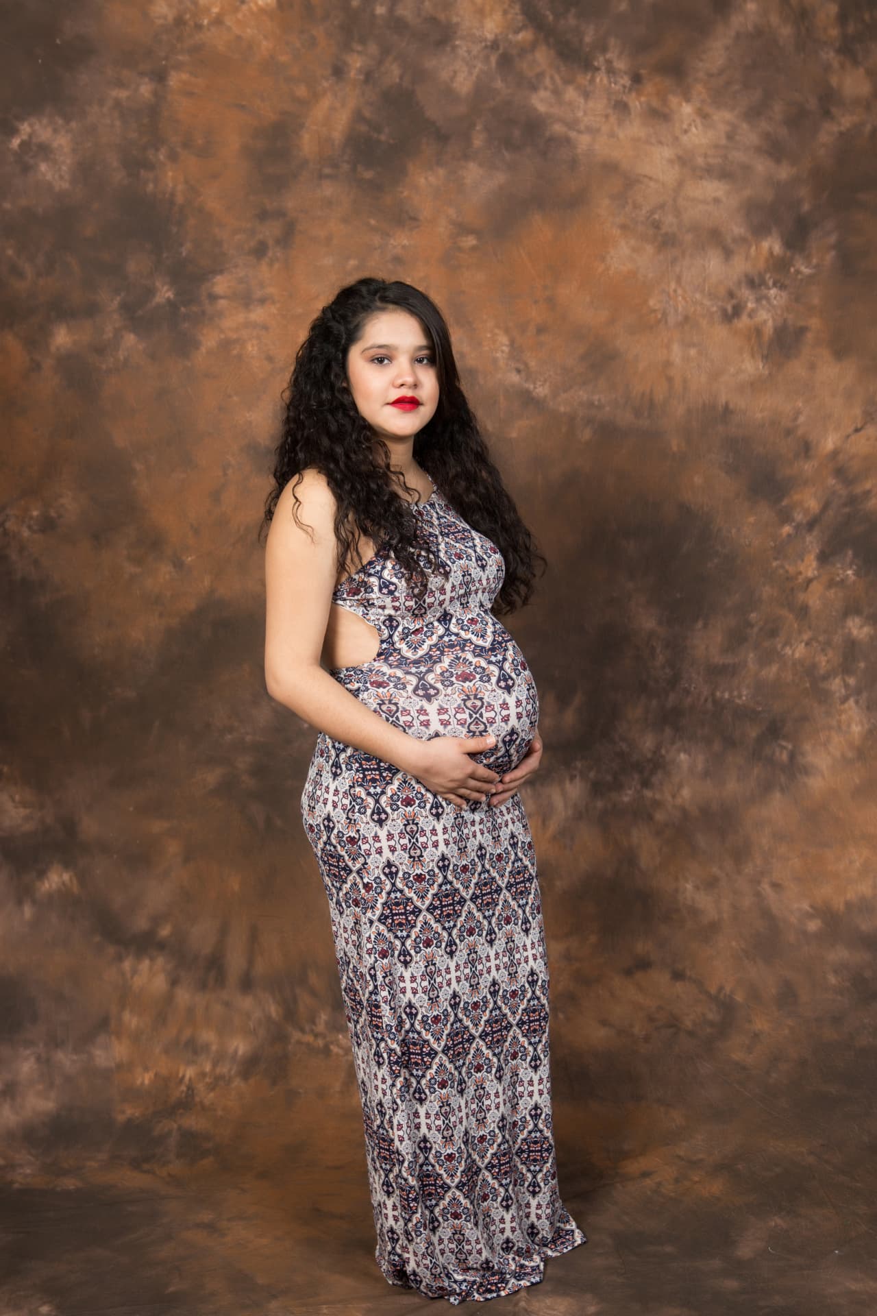 Expectant mother in a patterned gown posing before a warm studio backdrop