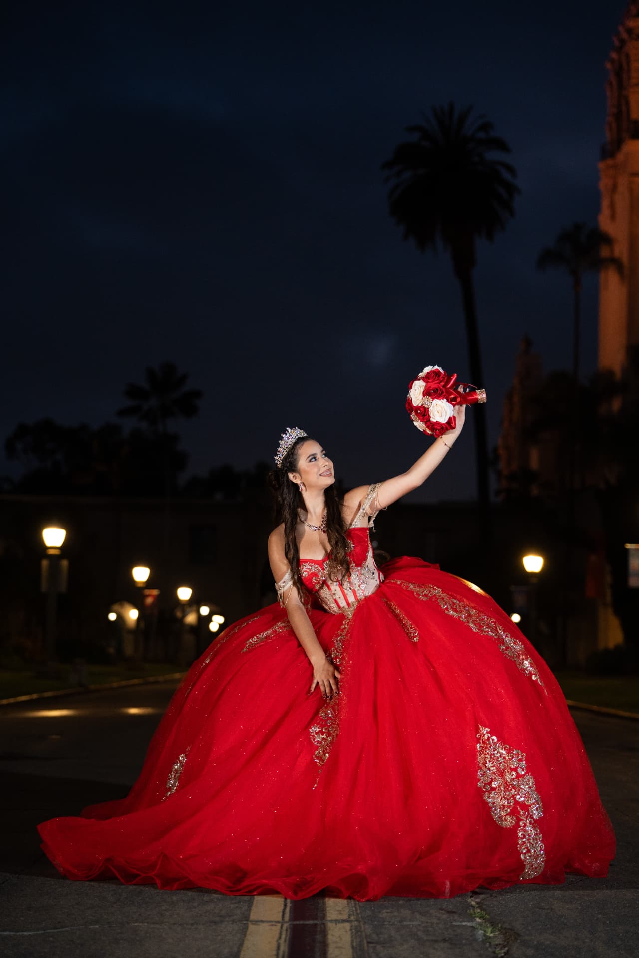 Quinceañera in a red gown raising her bouquet toward the night sky