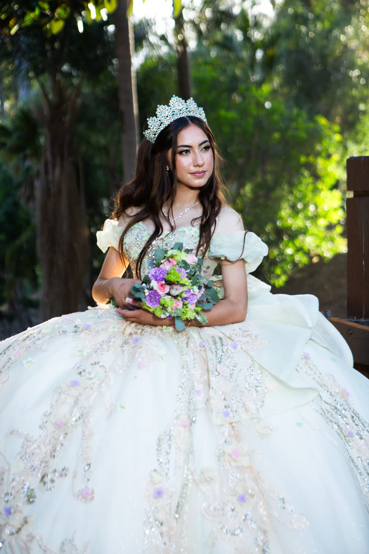 Quinceañera in an ornate cream gown holding a pastel bouquet in a garden