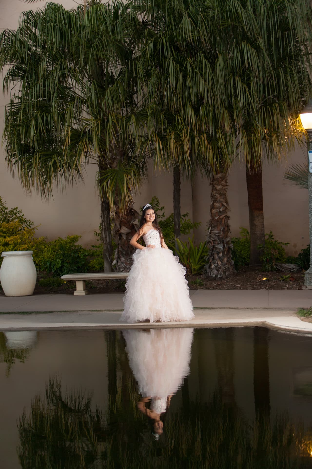 Quinceañera in a white gown reflected in a pond beneath palms at dusk