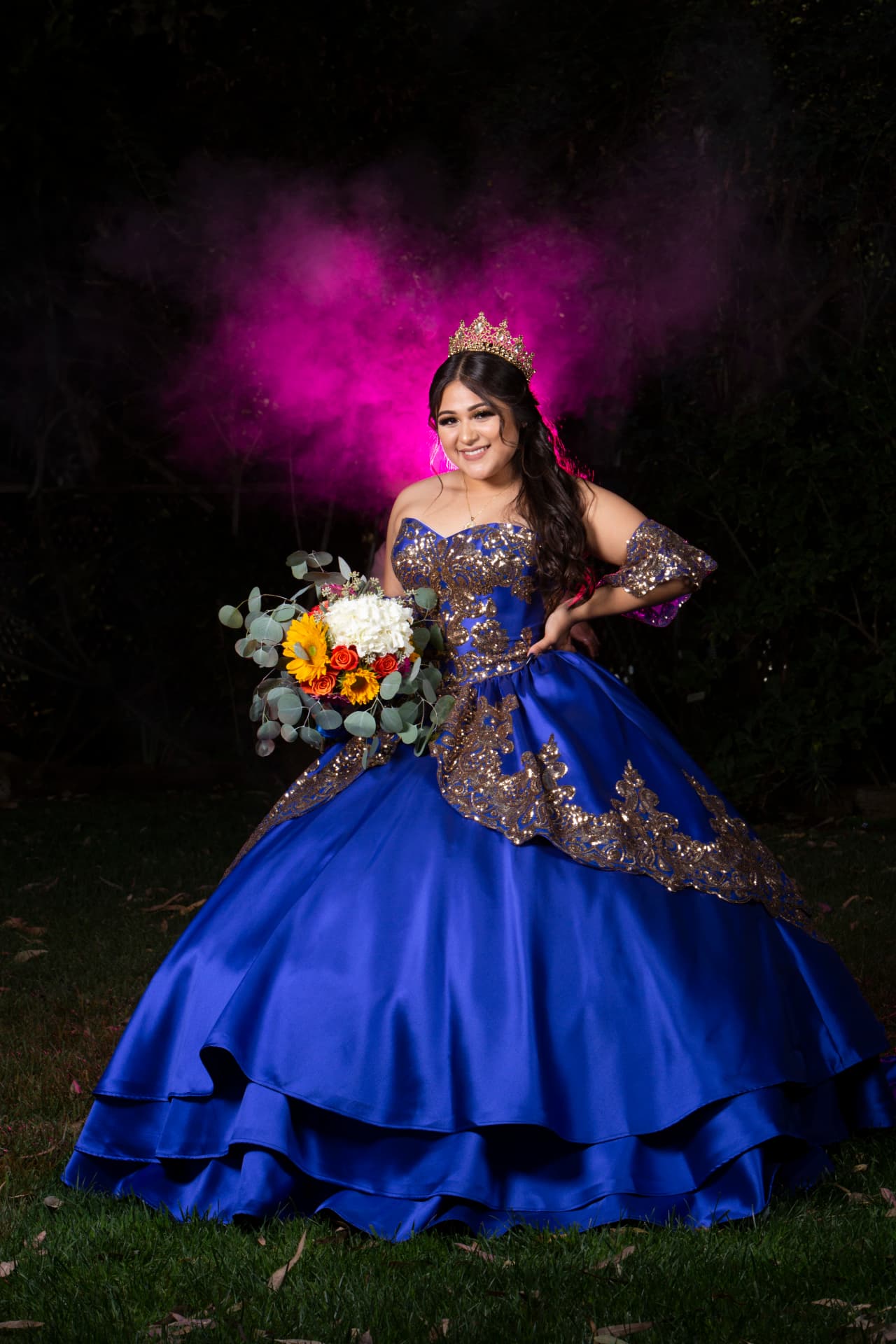 Quinceañera in a royal blue and gold gown with a sunflower bouquet against pink smoke at night