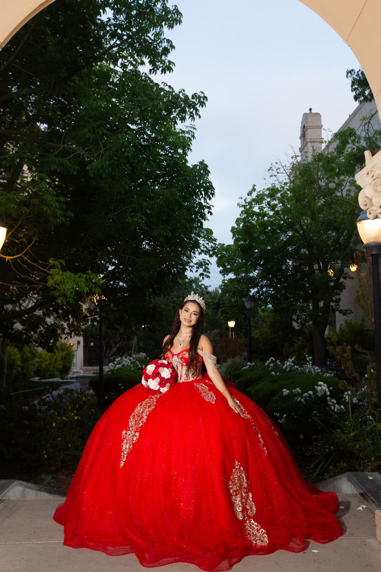 Quinceañera in a red gown standing beneath an archway with her bouquet at twilight