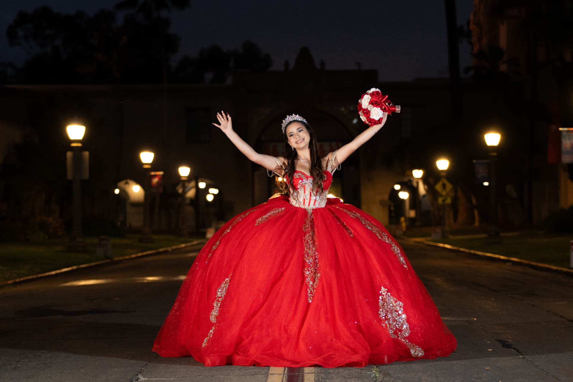 Quinceañera in a red gown with her arms outstretched on a lamp-lit road at night