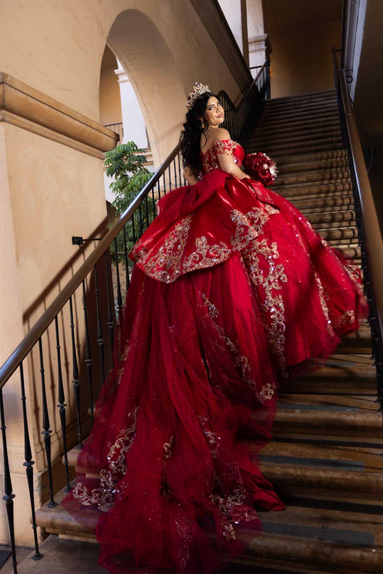 Quinceañera in a red and gold gown with her long train sweeping down a grand staircase
