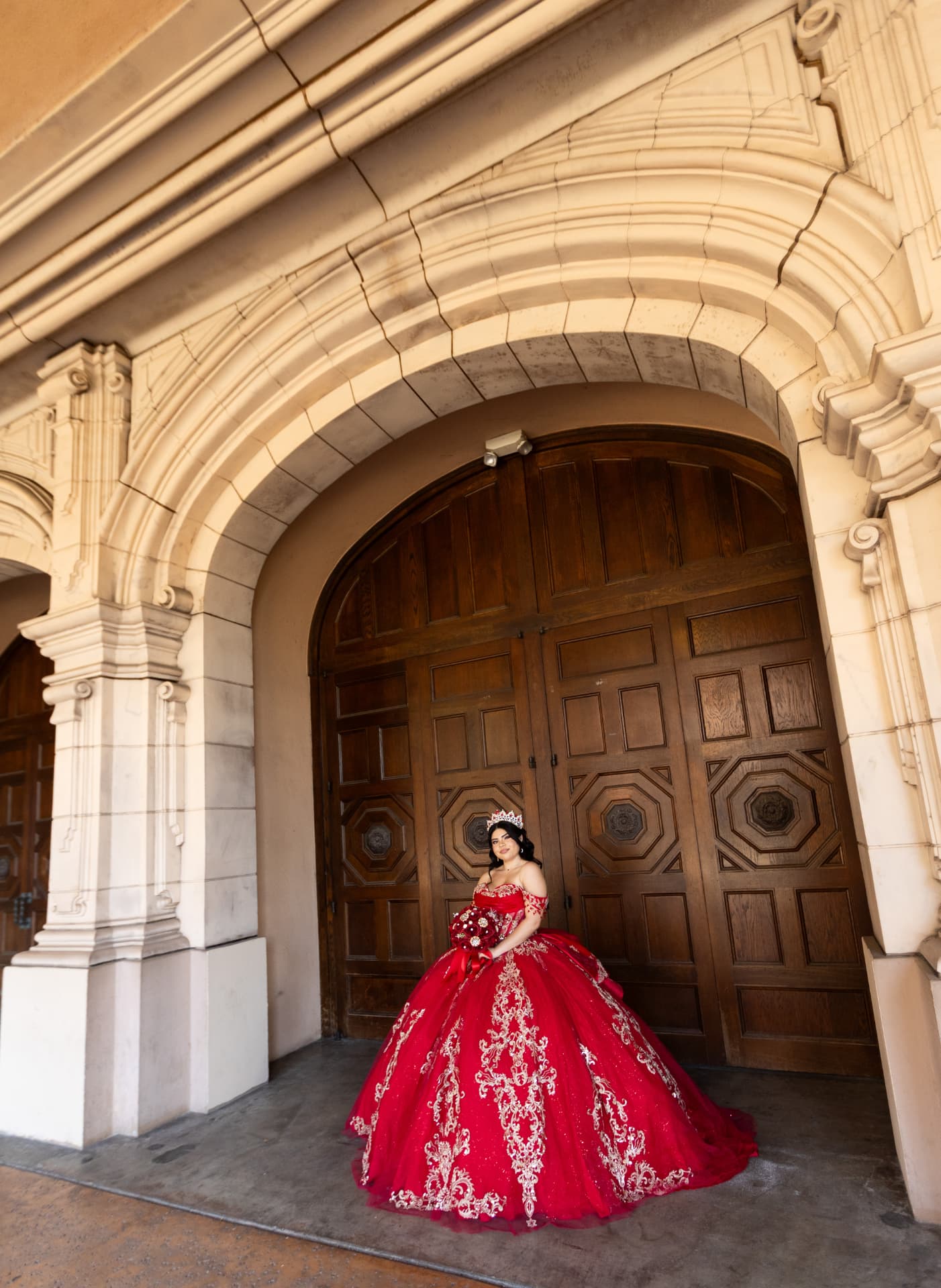 Quinceañera in a red and gold gown seated beneath a carved stone archway with ornate wooden doors