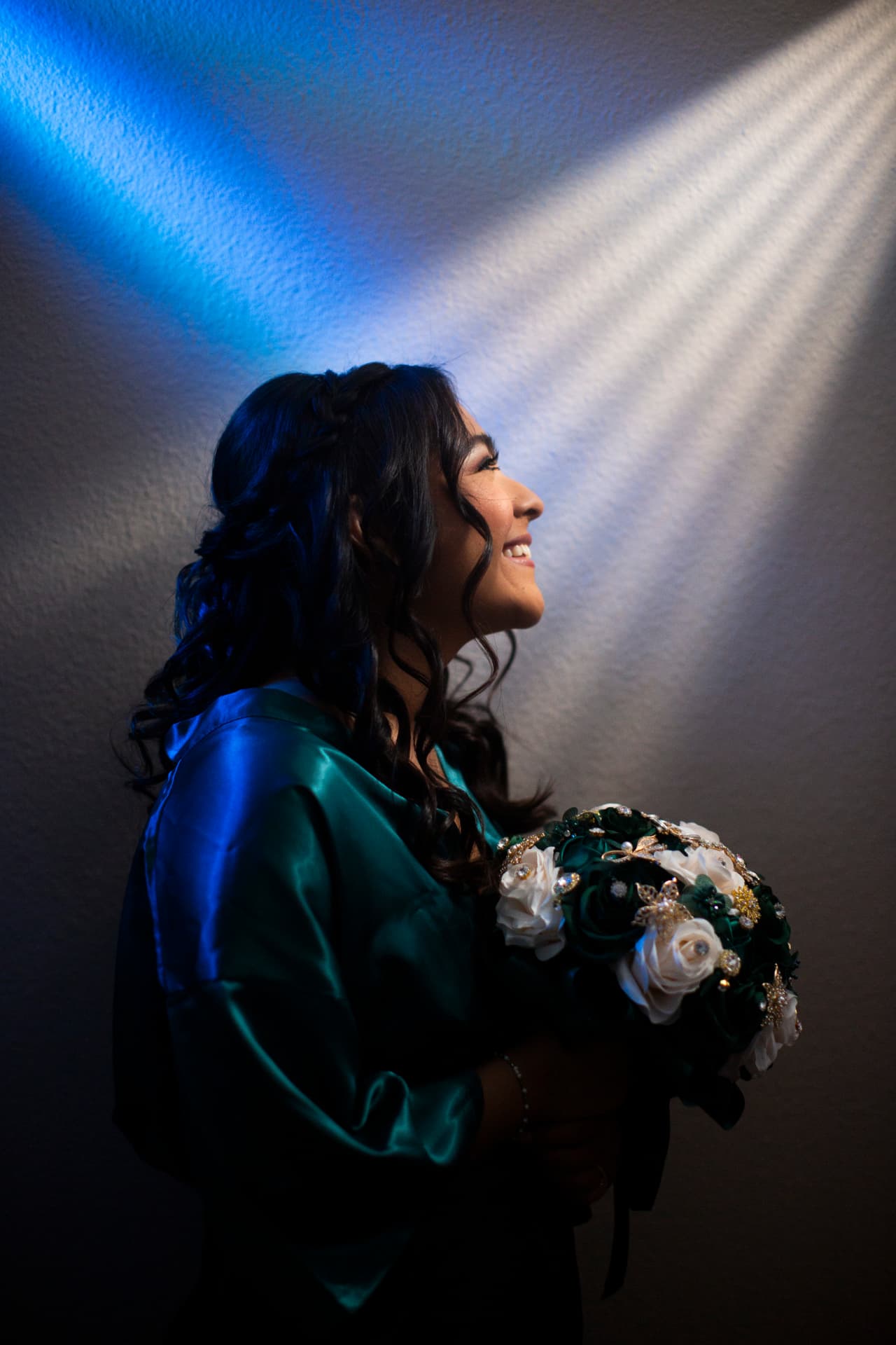 Quinceañera in an emerald satin robe laughing in blue and window light while holding her bouquet