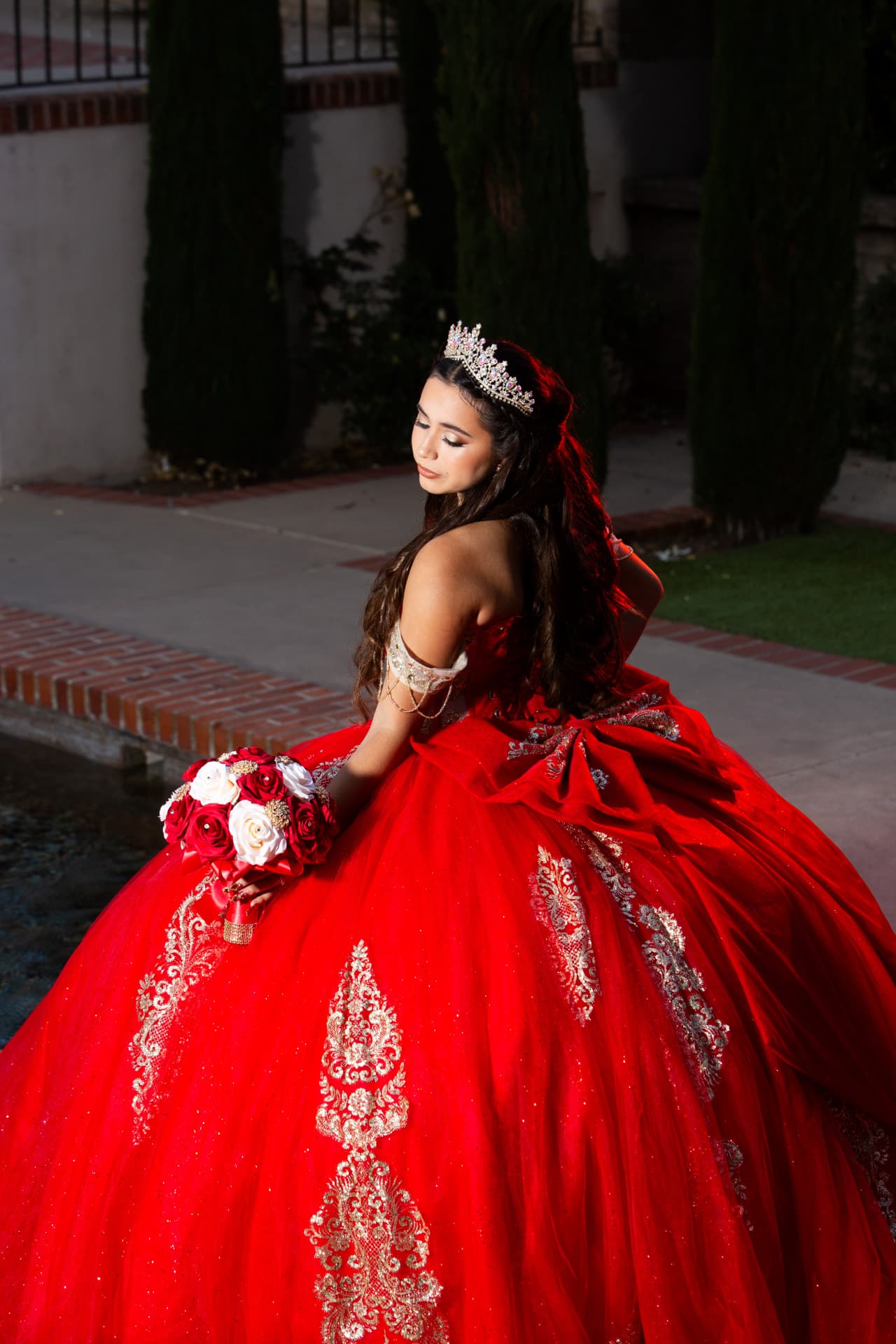 Quinceañera in a red and gold gown with a large bow, looking over her shoulder at dusk