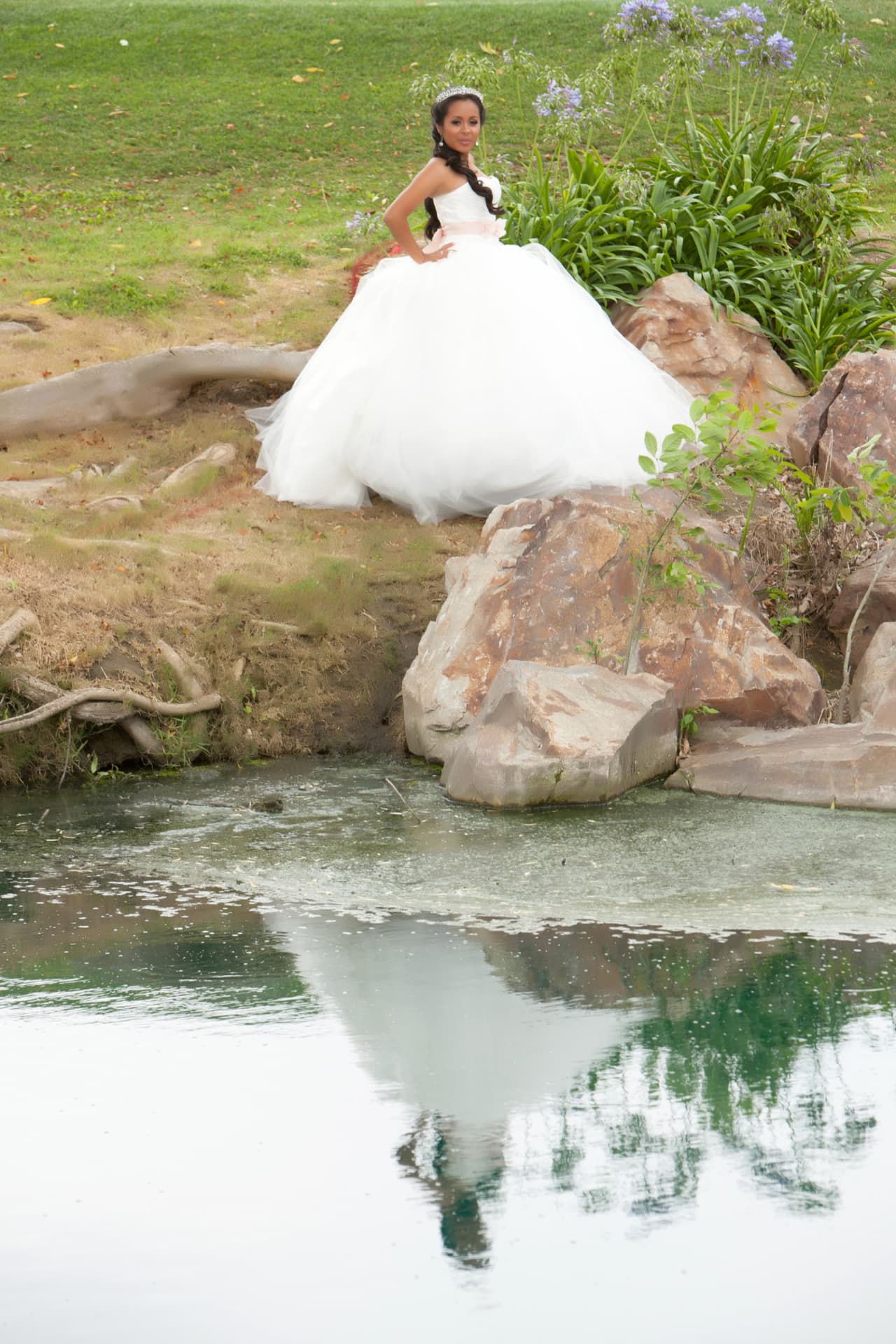 Quinceañera in a white gown and tiara seated on rocks above a still pond with purple flowers