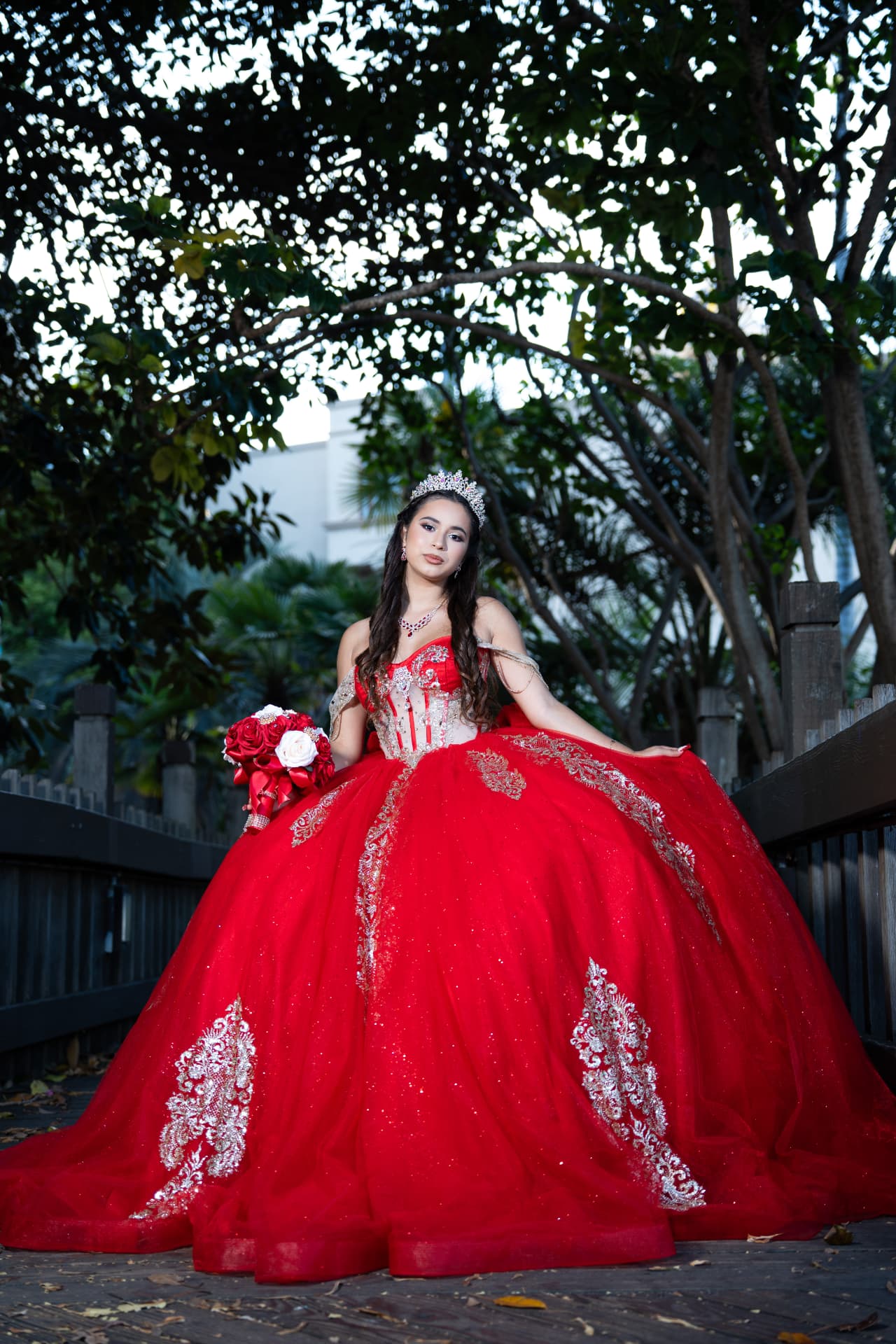 Quinceañera in a red gown with golden embroidery holding her bouquet on a garden path
