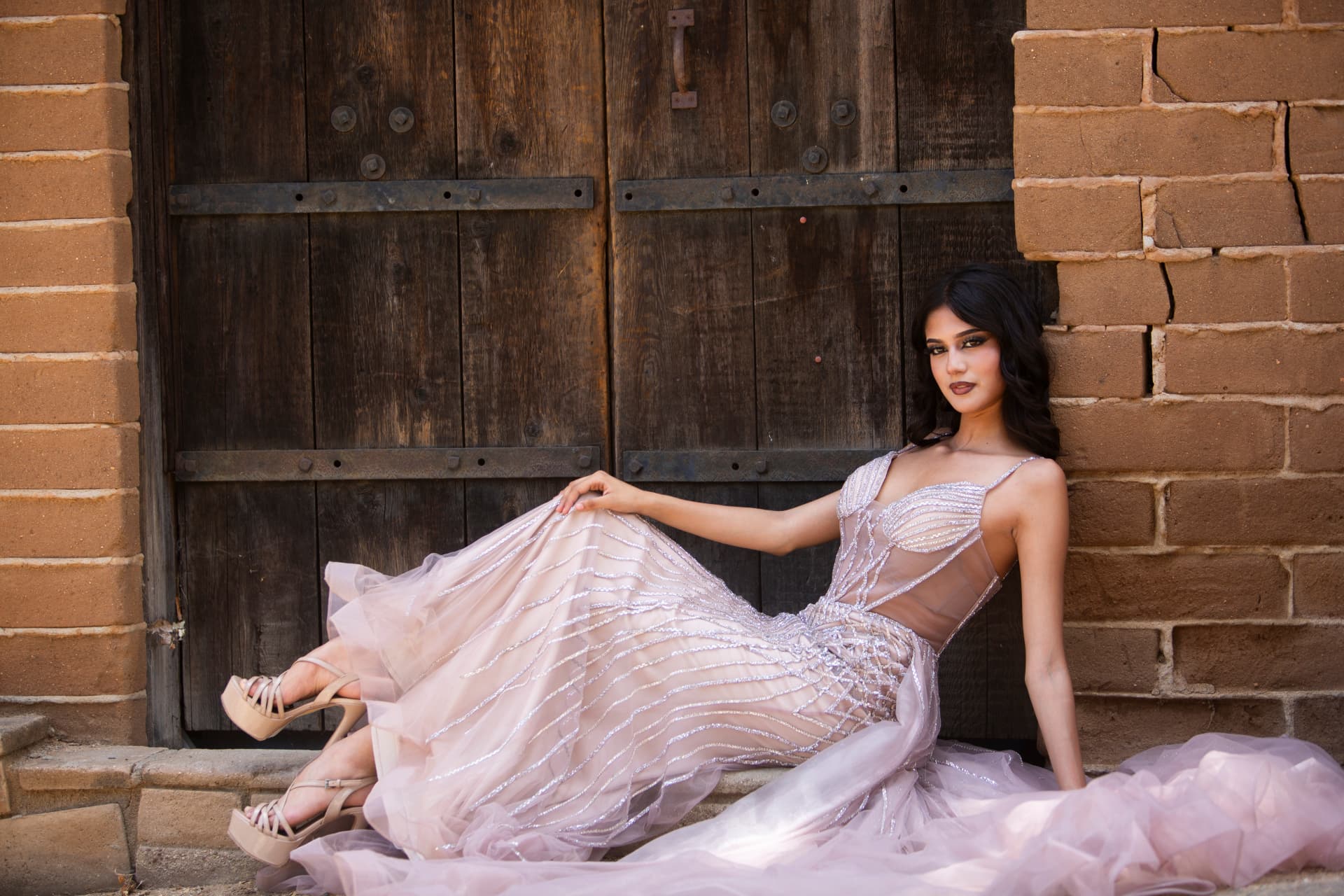 Fashion model reclining in a shimmering blush gown before rustic wooden doors