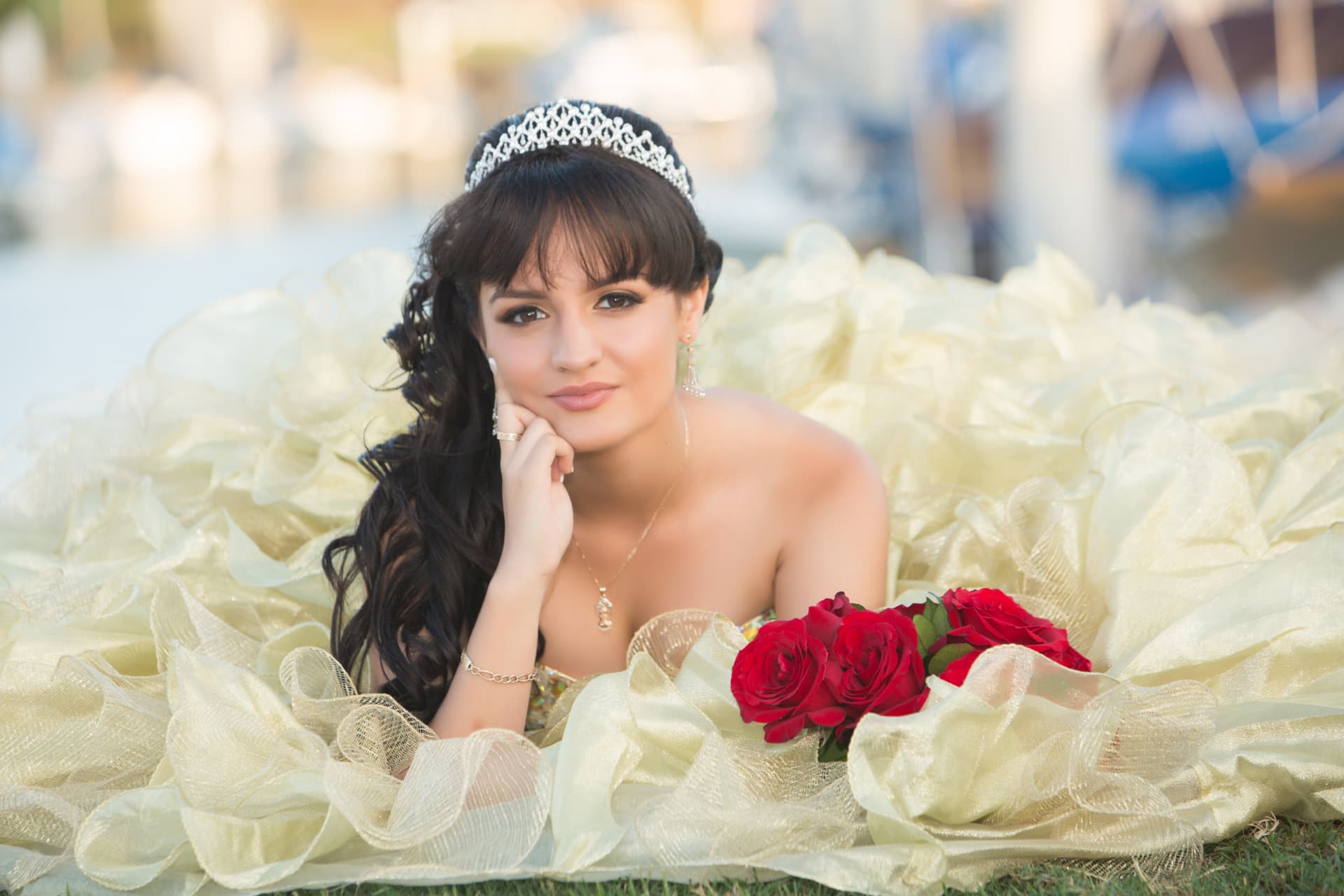 Quinceañera in a gold gown lying on the grass with red roses, chin resting on her hand