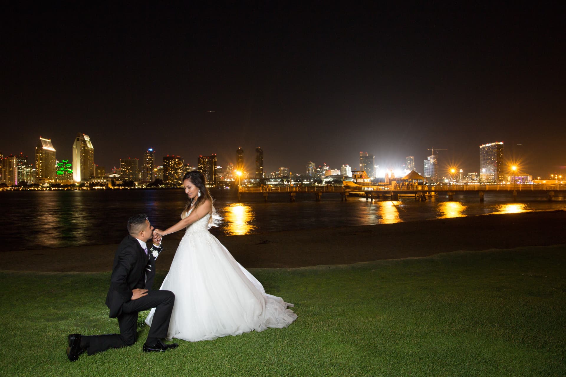 Groom kneeling to kiss his bride’s hand before a luminous waterfront skyline
