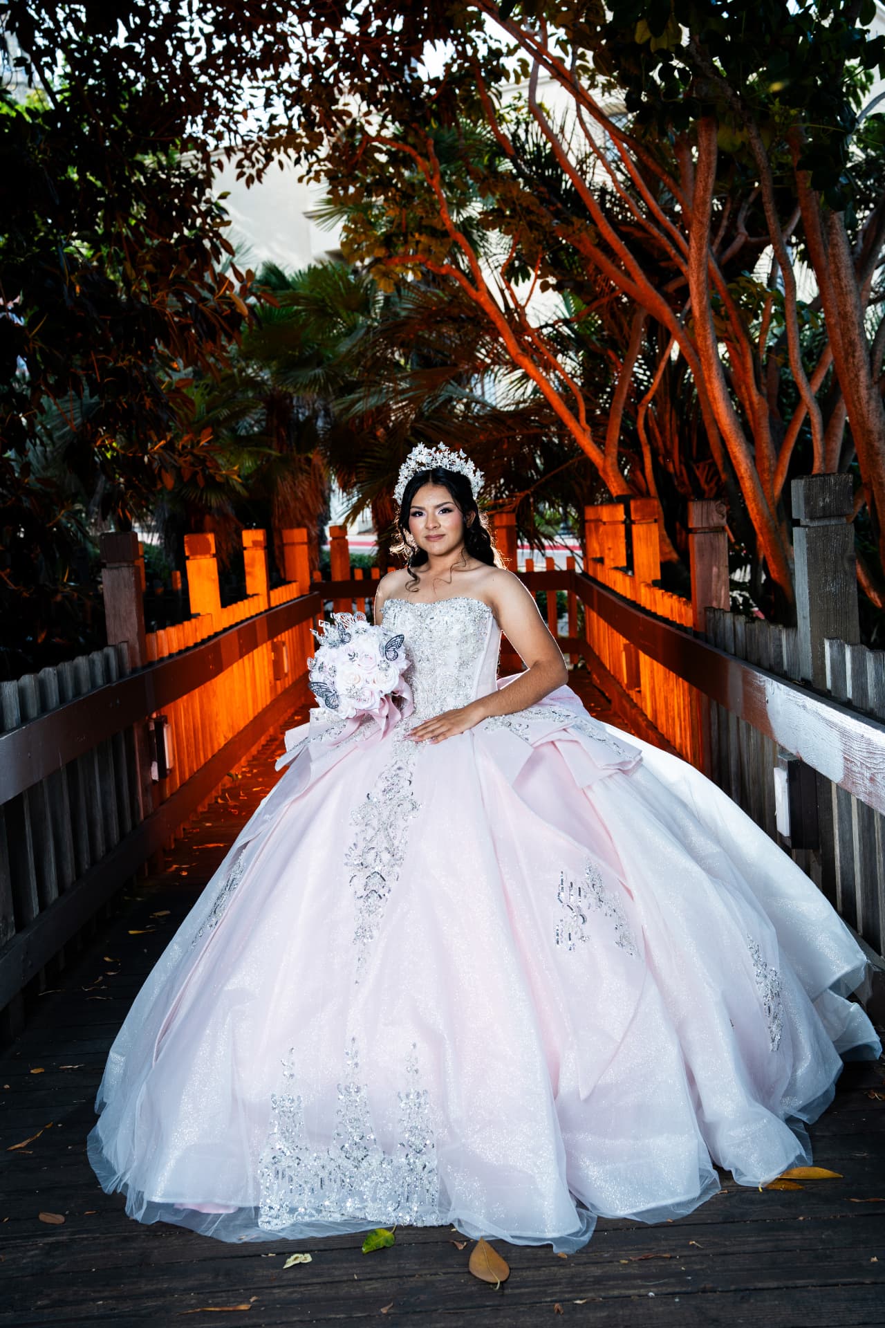 Quinceañera in a pink gown with a butterfly bouquet on a warmly lit wooden bridge at dusk