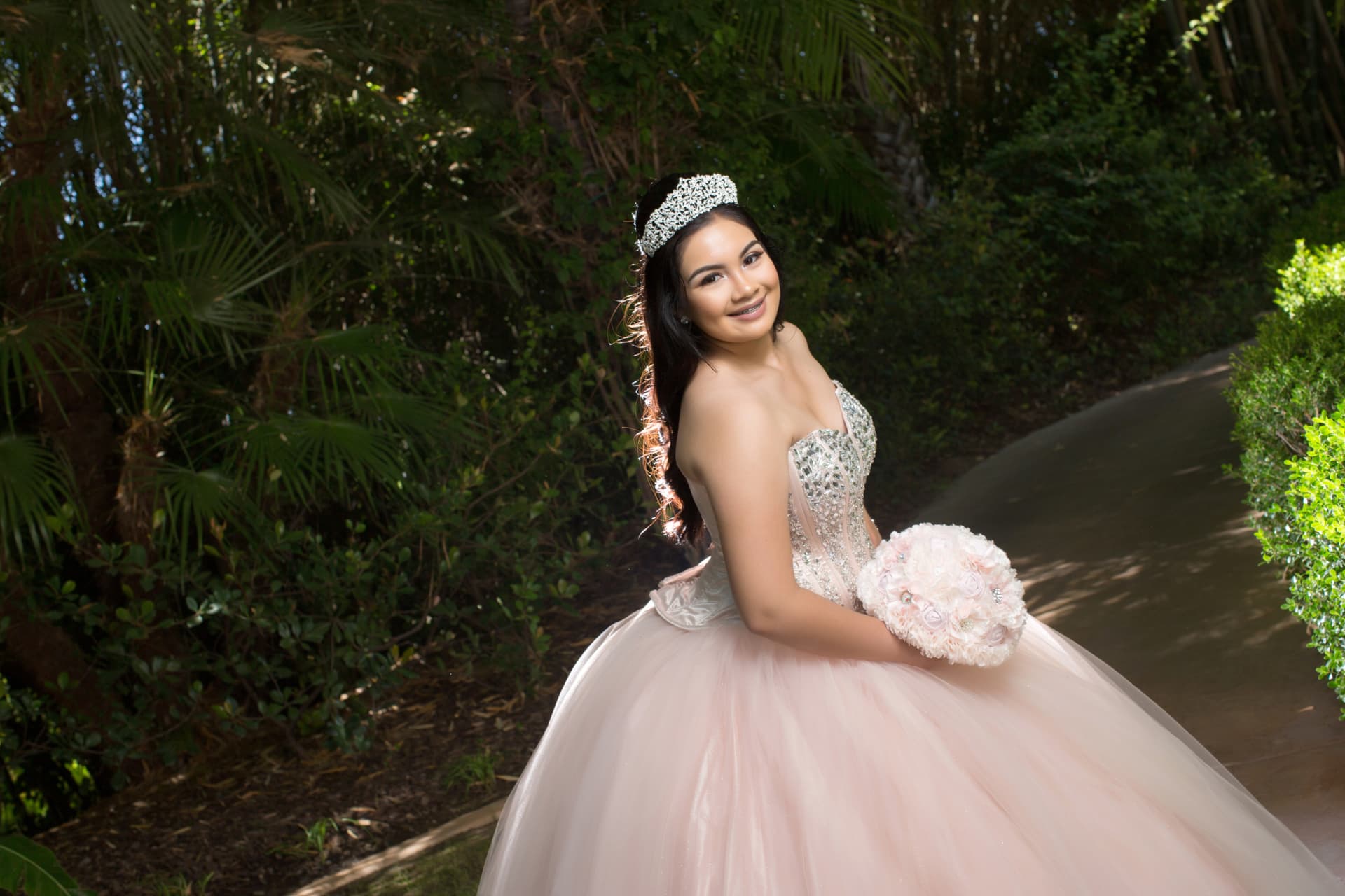 Quinceañera in a blush gown and sparkling tiara holding a pink bouquet on a garden path