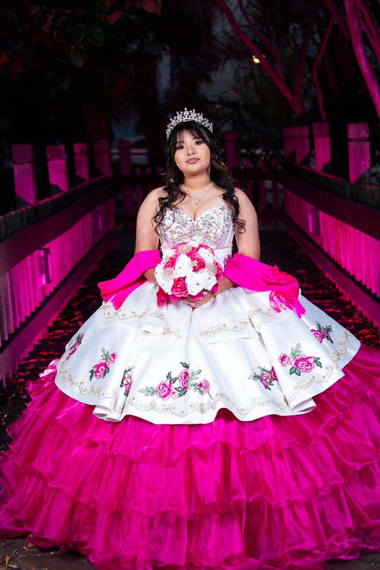 Quinceañera in a white and pink gown seated on a bridge lit pink at night