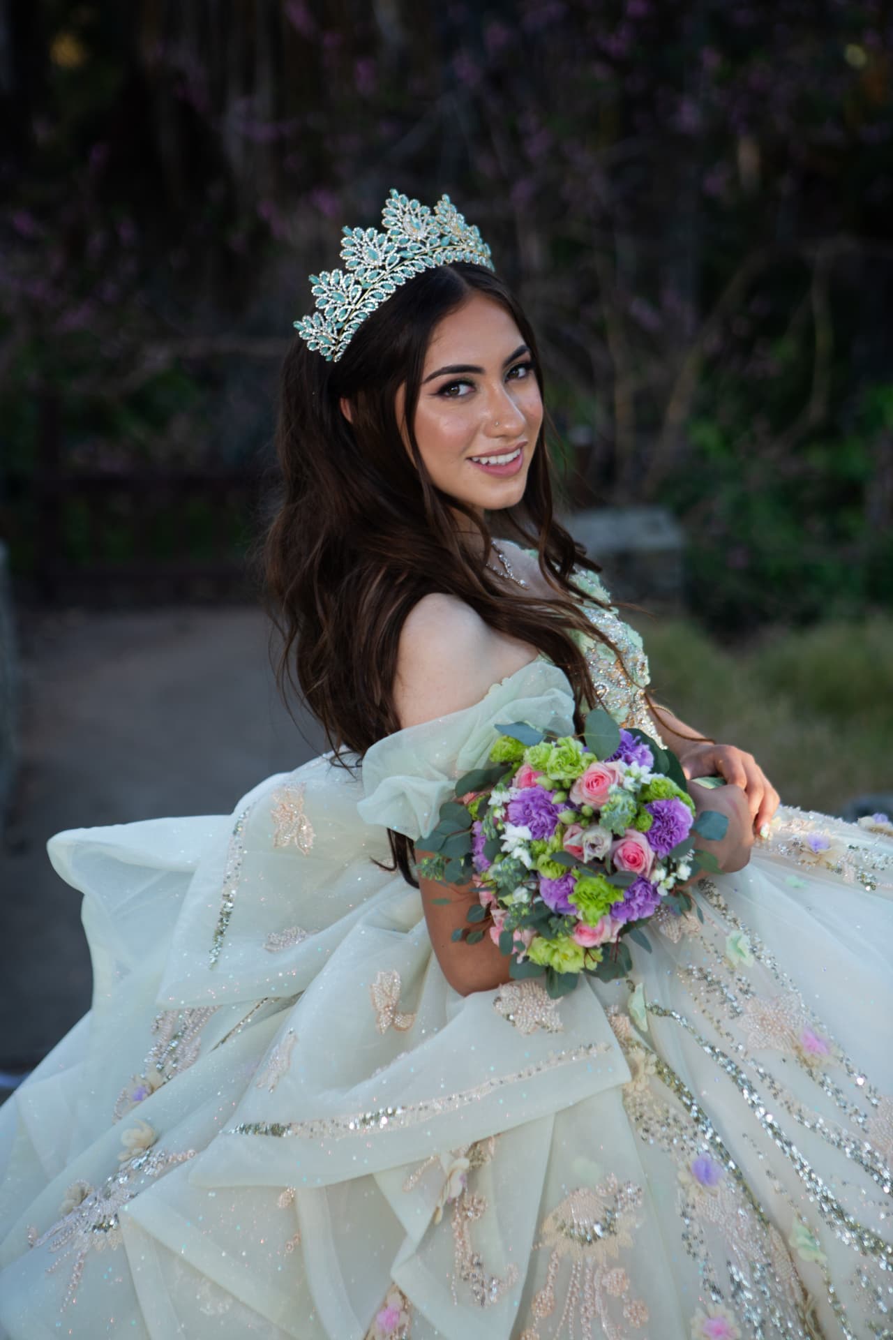 Smiling quinceañera with a jeweled crown holding a colorful bouquet in a garden