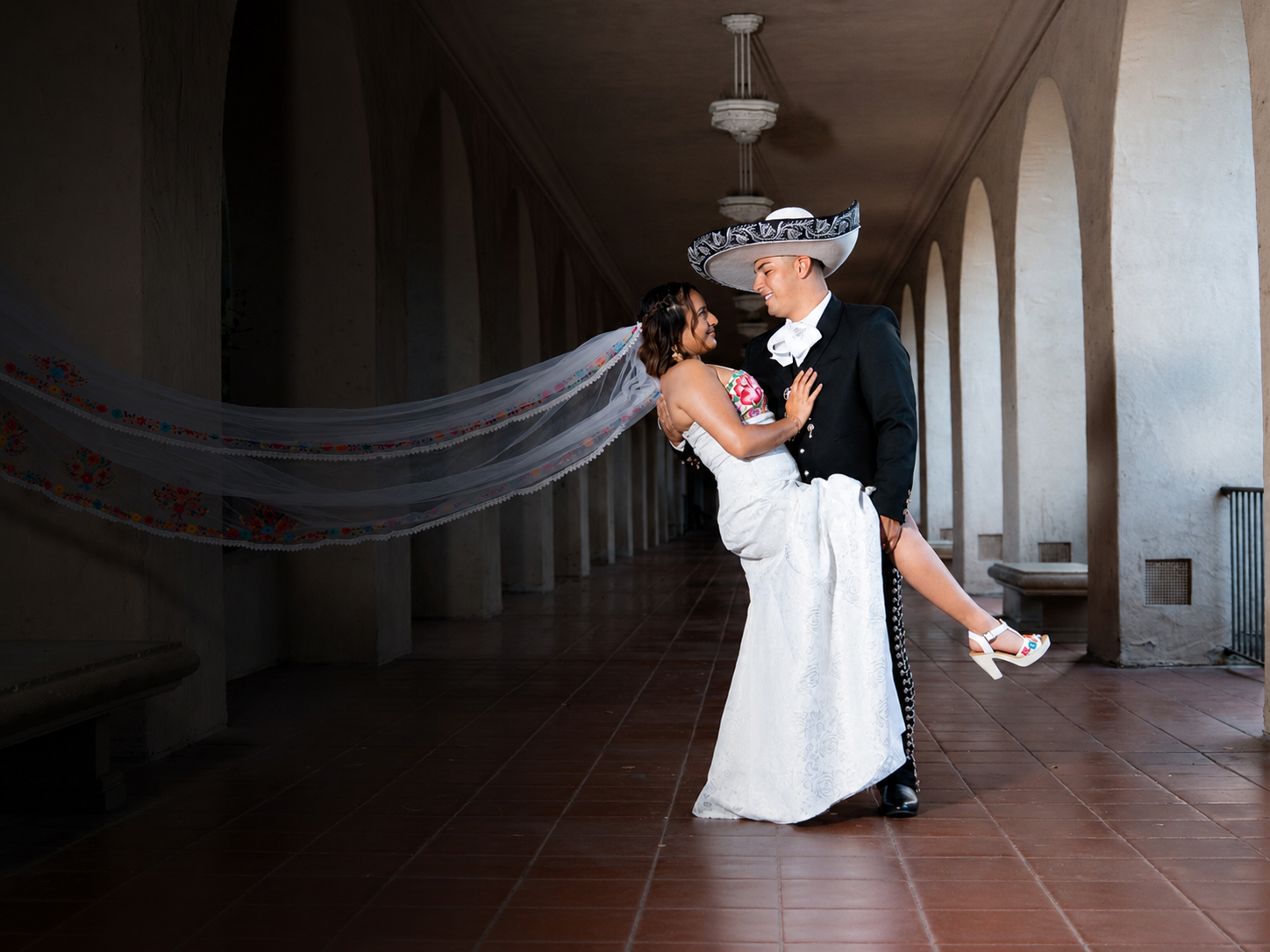 Groom in a sombrero de charro dips his bride beneath arches as her embroidered veil sweeps across the corridor