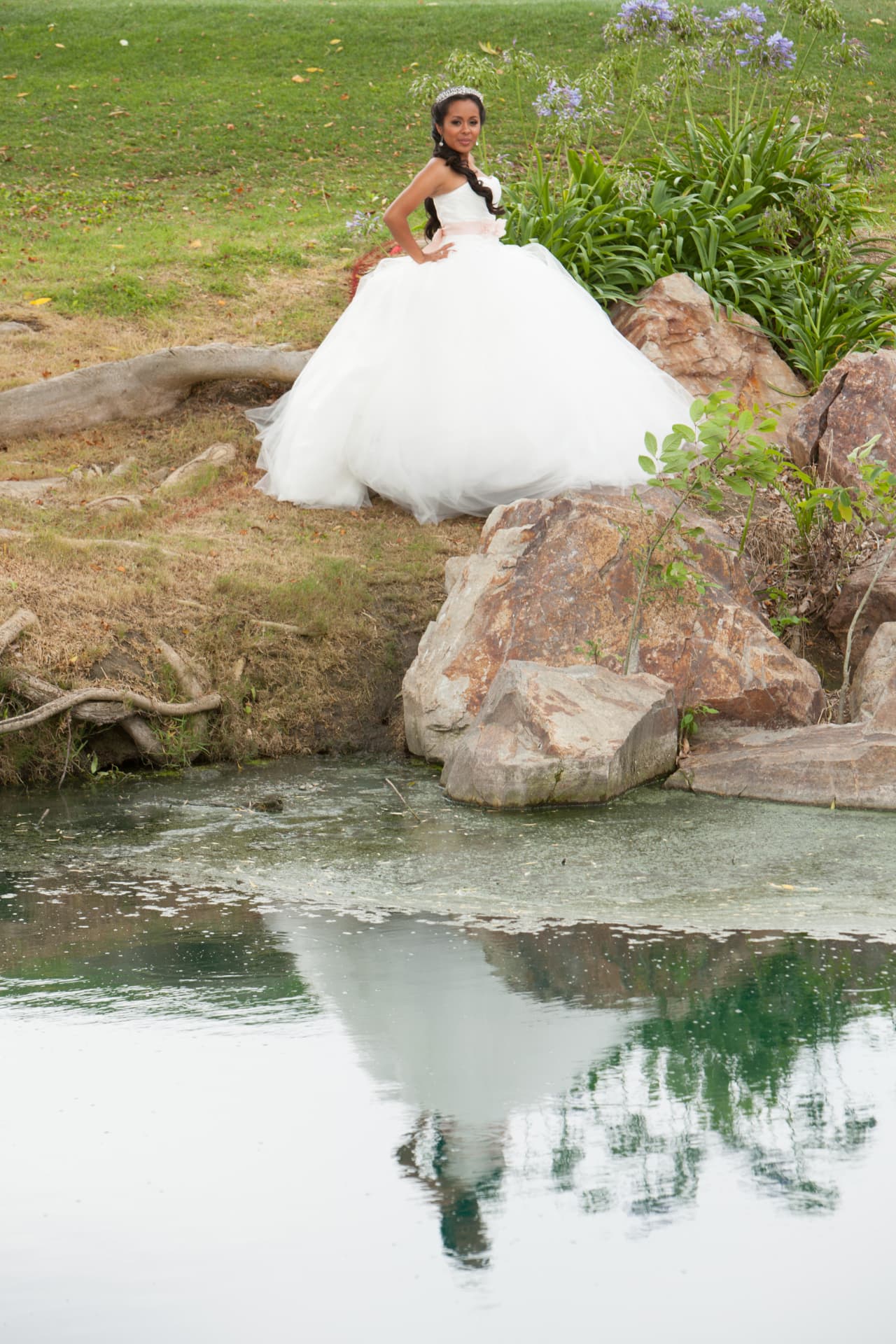 Quinceañera in a white gown reflected in a still pond beside rocks and agapanthus