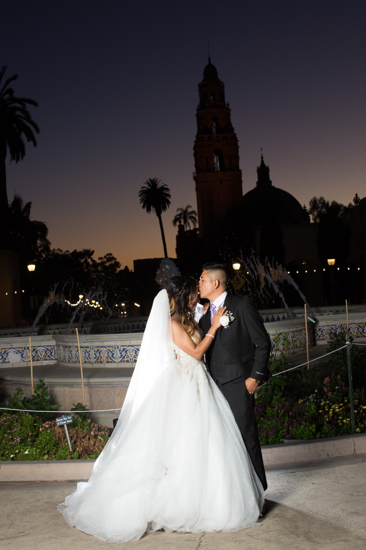 Bride and groom kissing beside a fountain beneath a historic tower at twilight