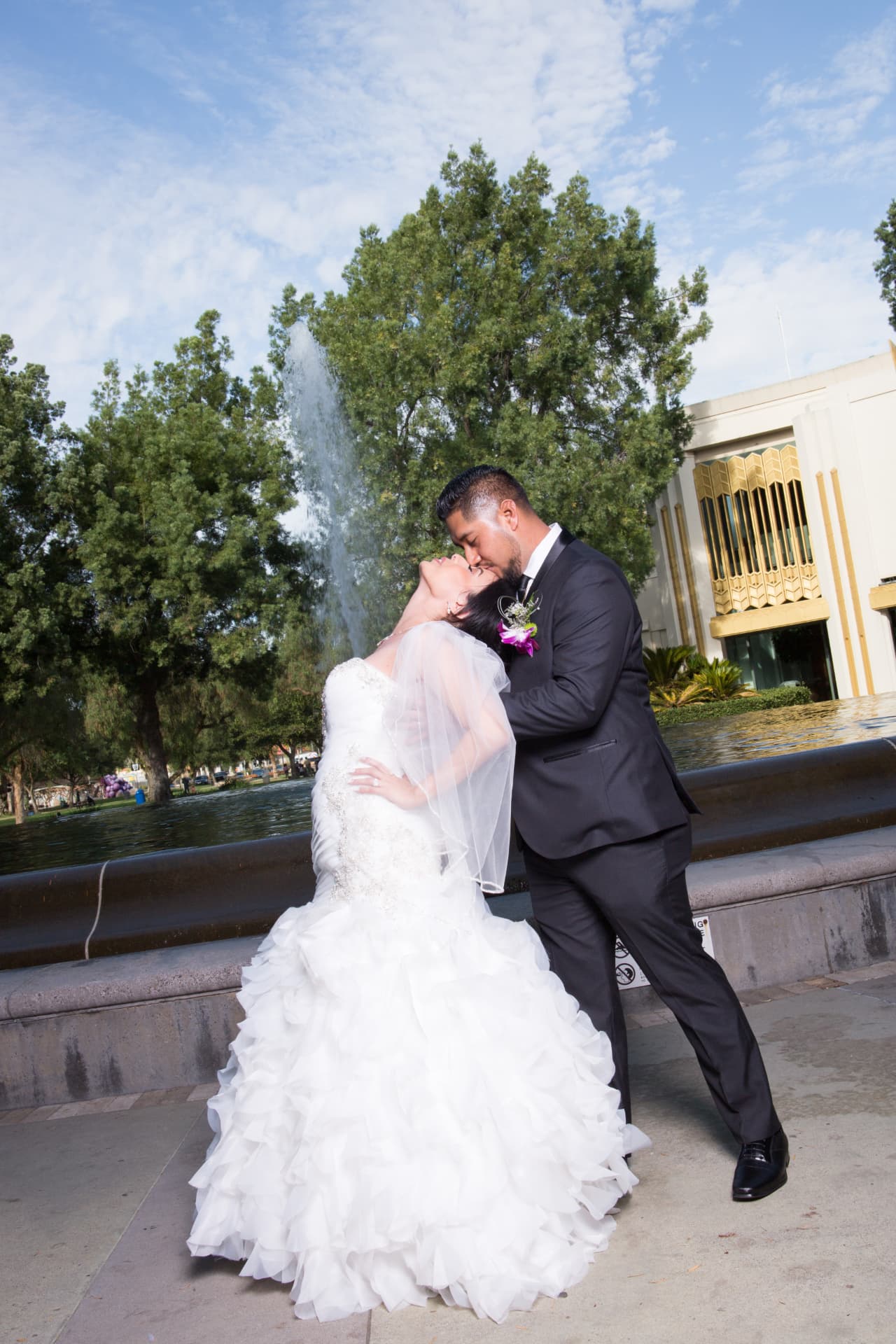 Groom kissing his bride beside a fountain beneath a bright blue sky