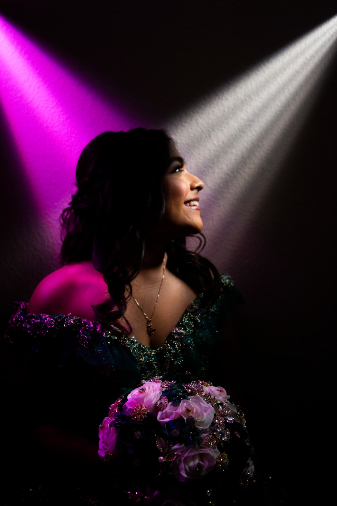 Quinceañera in an emerald gown glowing under magenta light with a jeweled bouquet