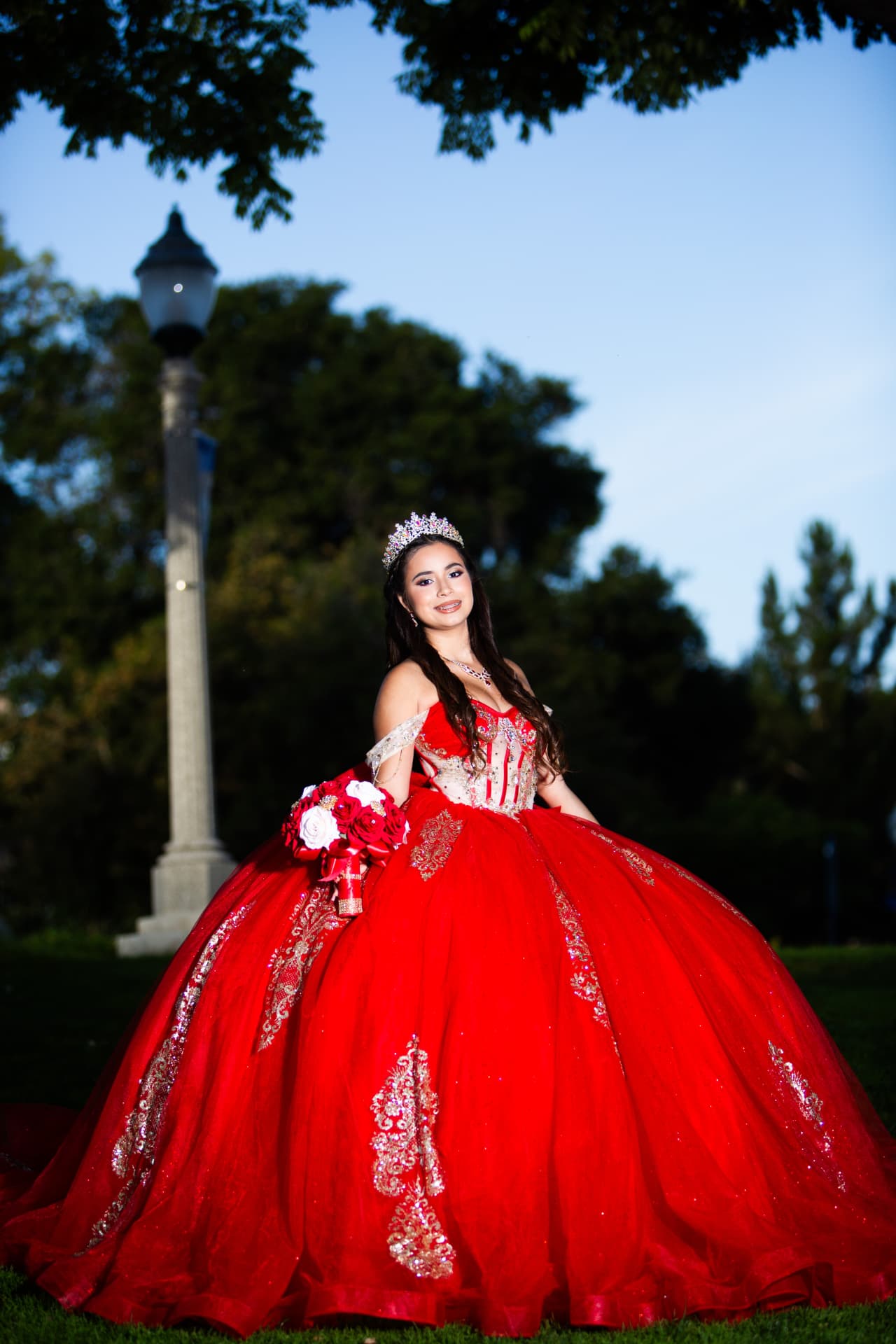 Quinceañera in a red and gold gown with her bouquet beside a lamp post in the park