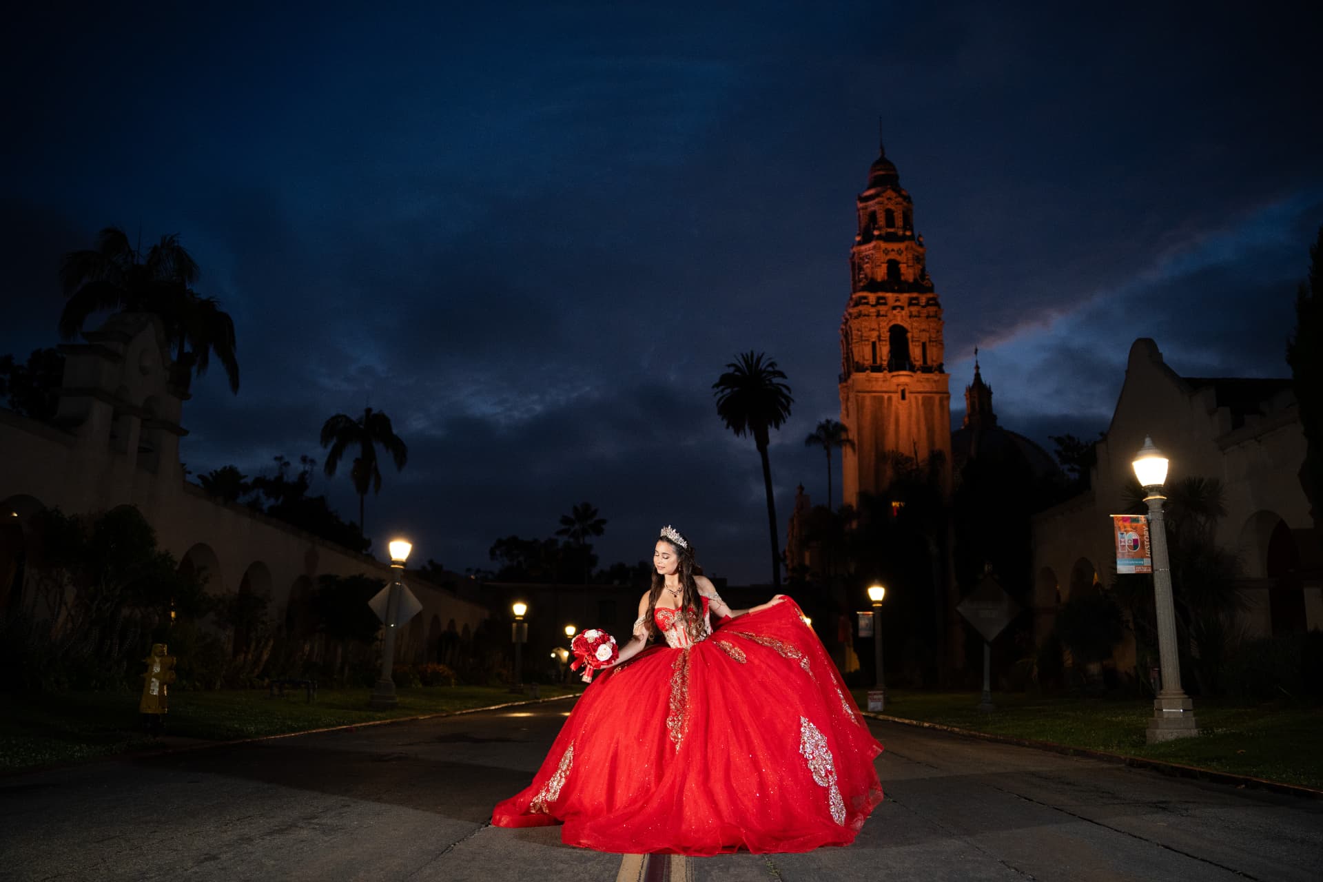Quinceañera in a red gown on a lamp-lit street with the Balboa Park tower lit at twilight