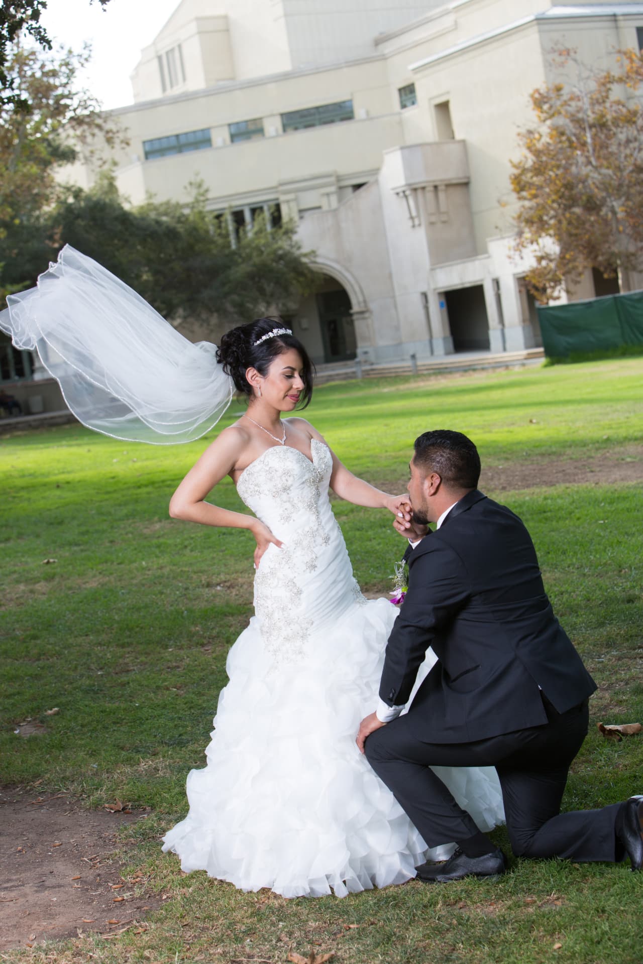 Groom kneeling to kiss his bride’s hand as her veil floats in the breeze
