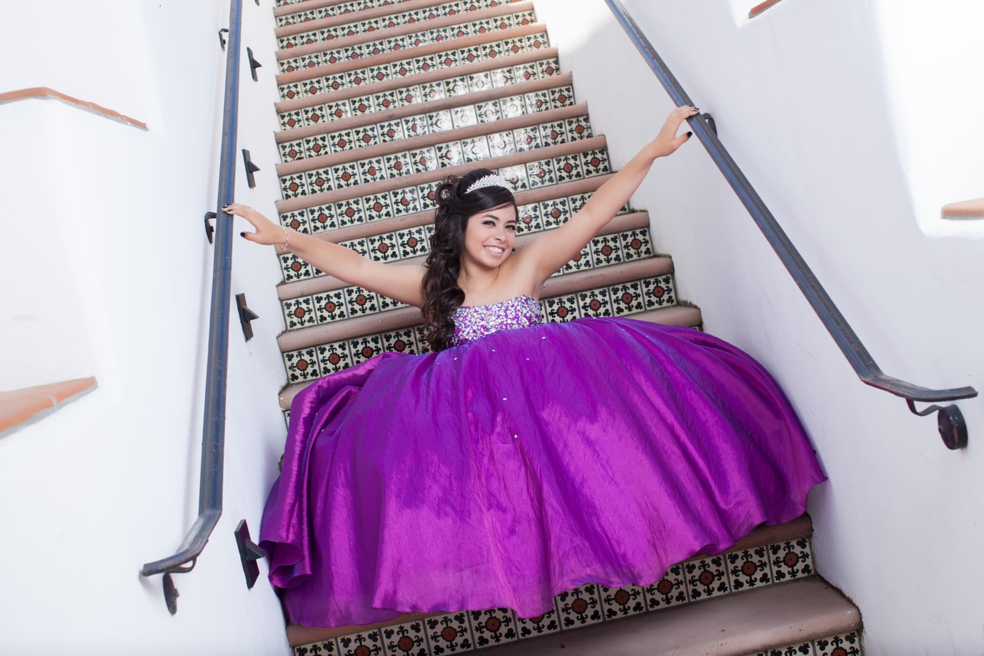 Quinceañera in a vivid purple gown stretching her arms wide on Spanish-tile stairs