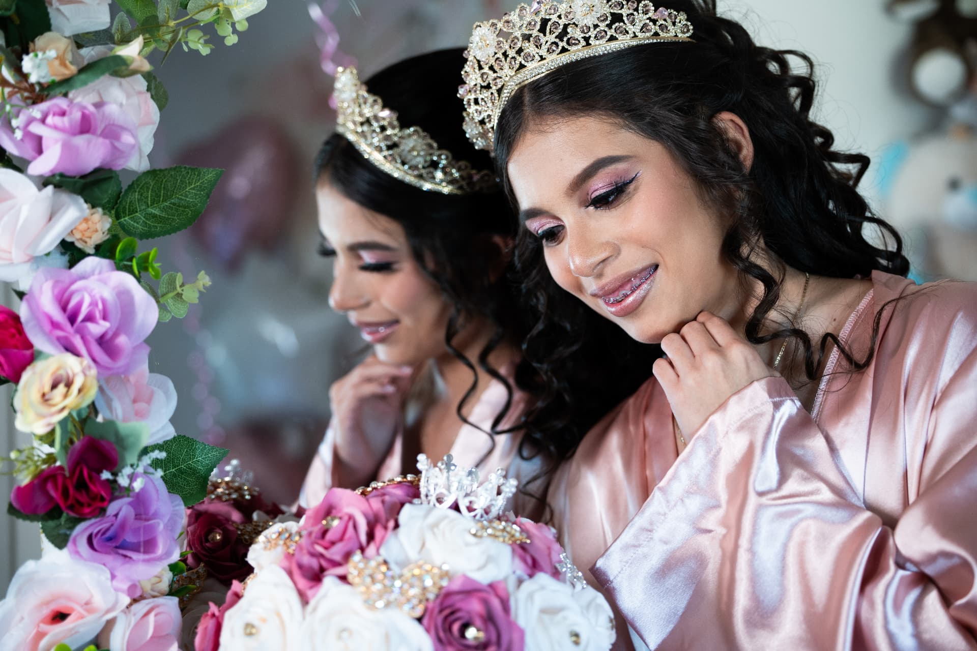 Quinceañera in a pink satin robe smiling beside the mirror before her quinceañera