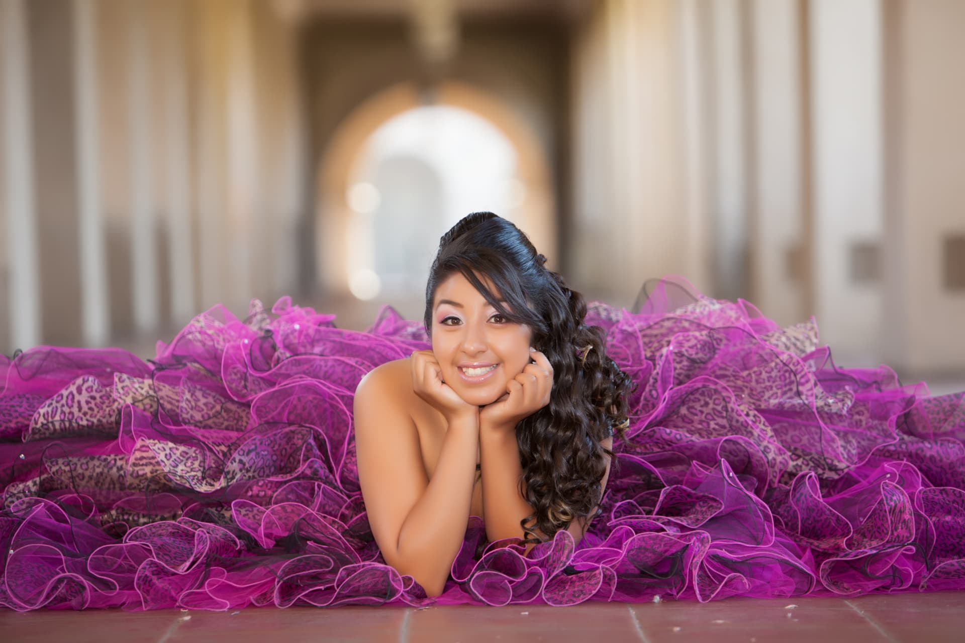 Quinceañera smiling in a magenta ruffled gown spread wide in an arched corridor