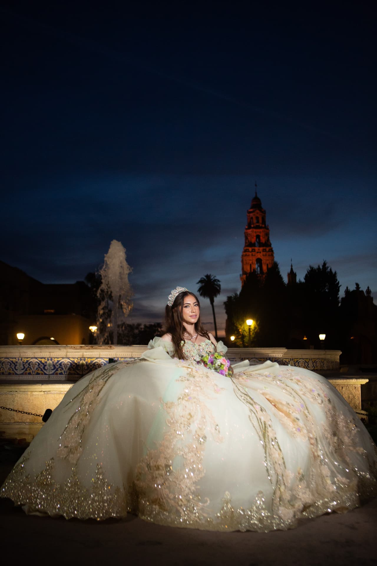Quinceañera in a sparkling gown seated before a fountain with the Balboa Park tower lit at twilight
