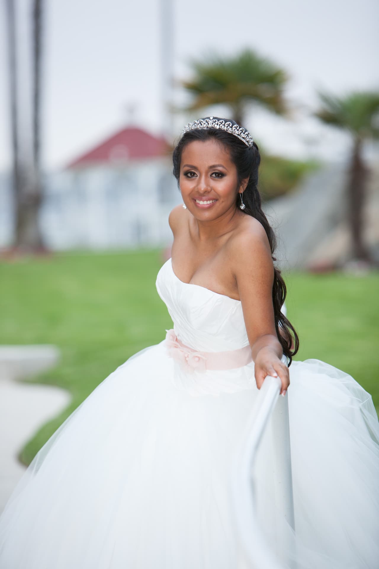 Quinceañera in a white gown smiling on a seaside lawn with palms and a gazebo behind her