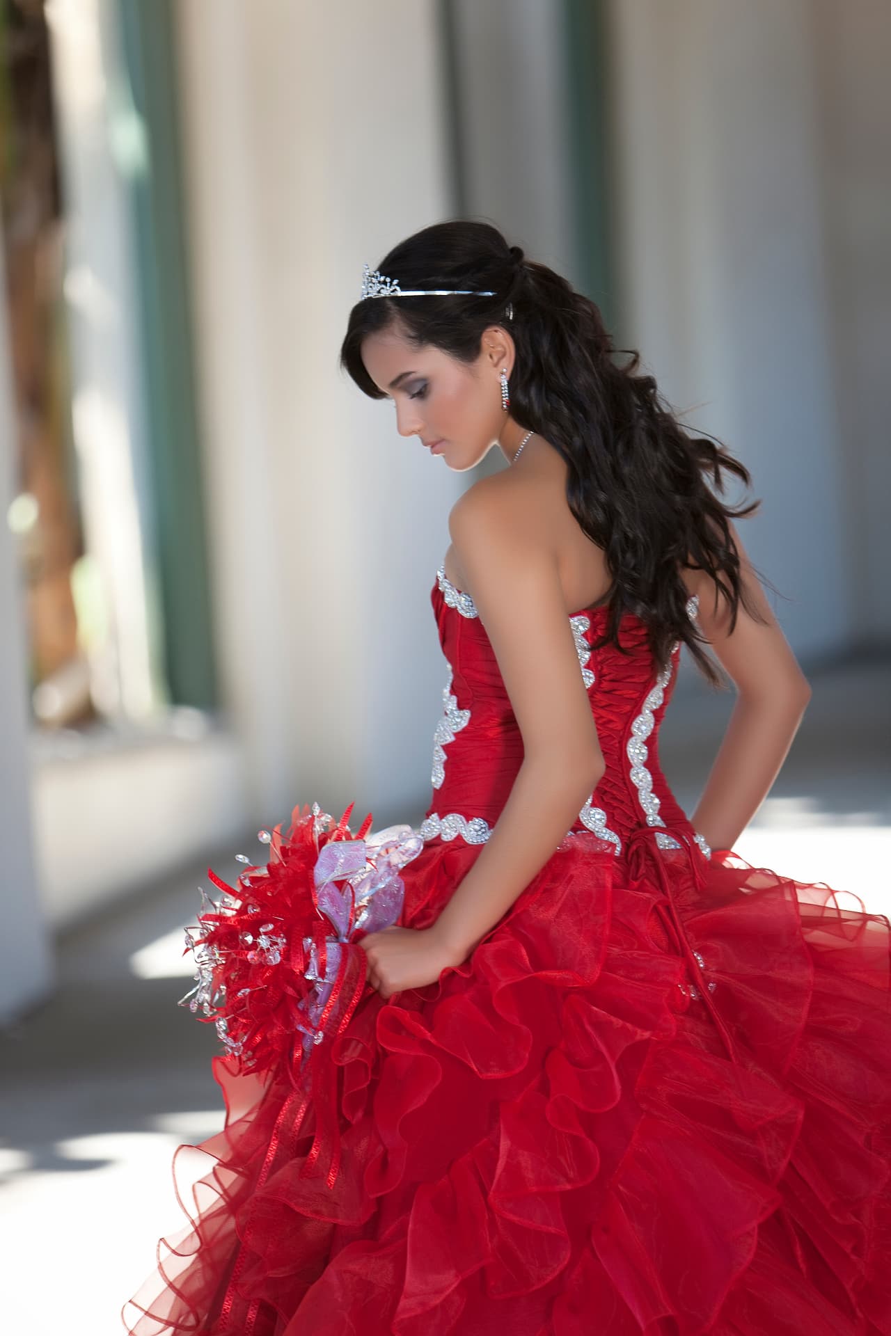 Quinceañera in a red ruffled gown looking down at her bouquet in an arched corridor