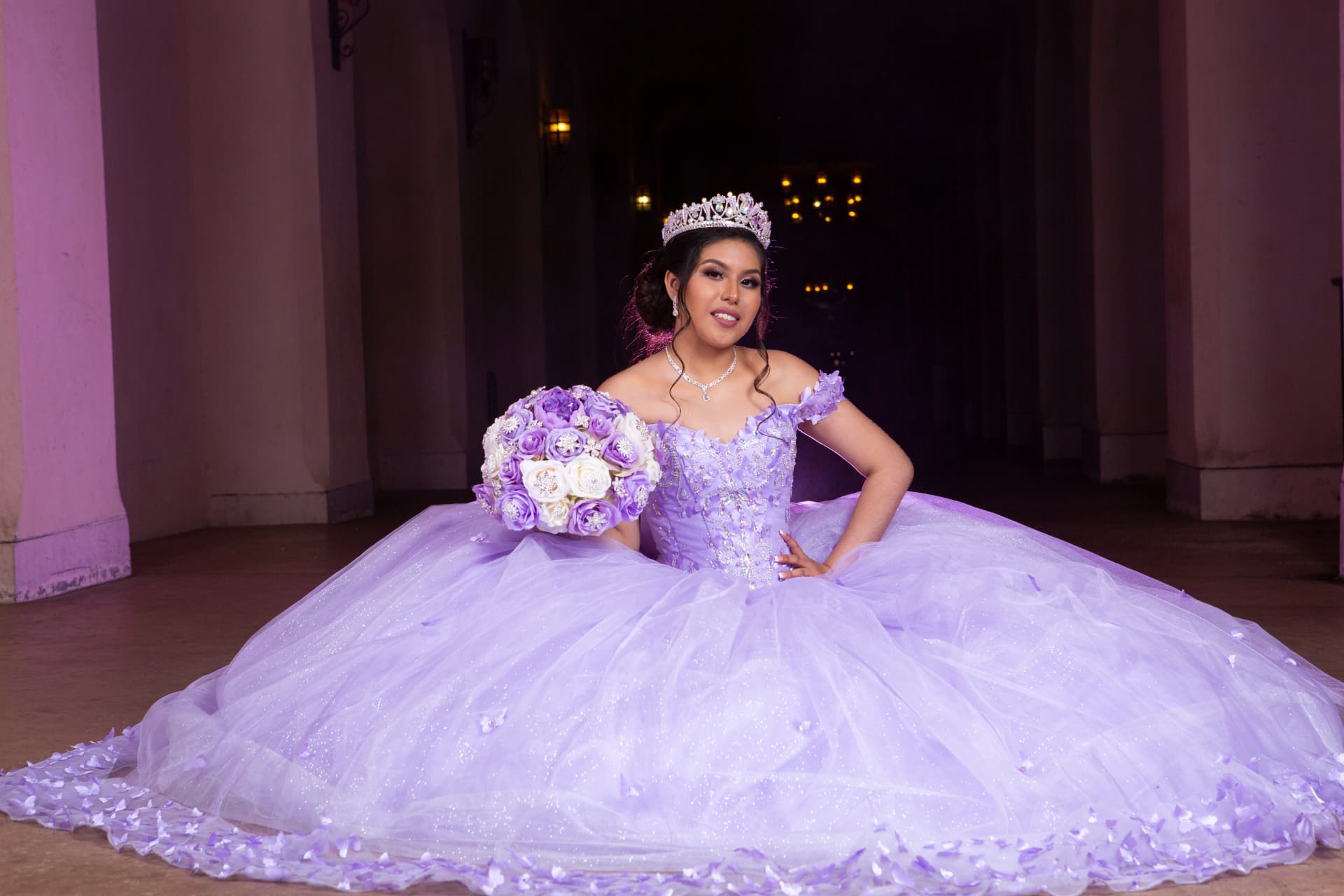 Quinceañera in a lavender gown holding a lavender and white bouquet beside purple-lit columns