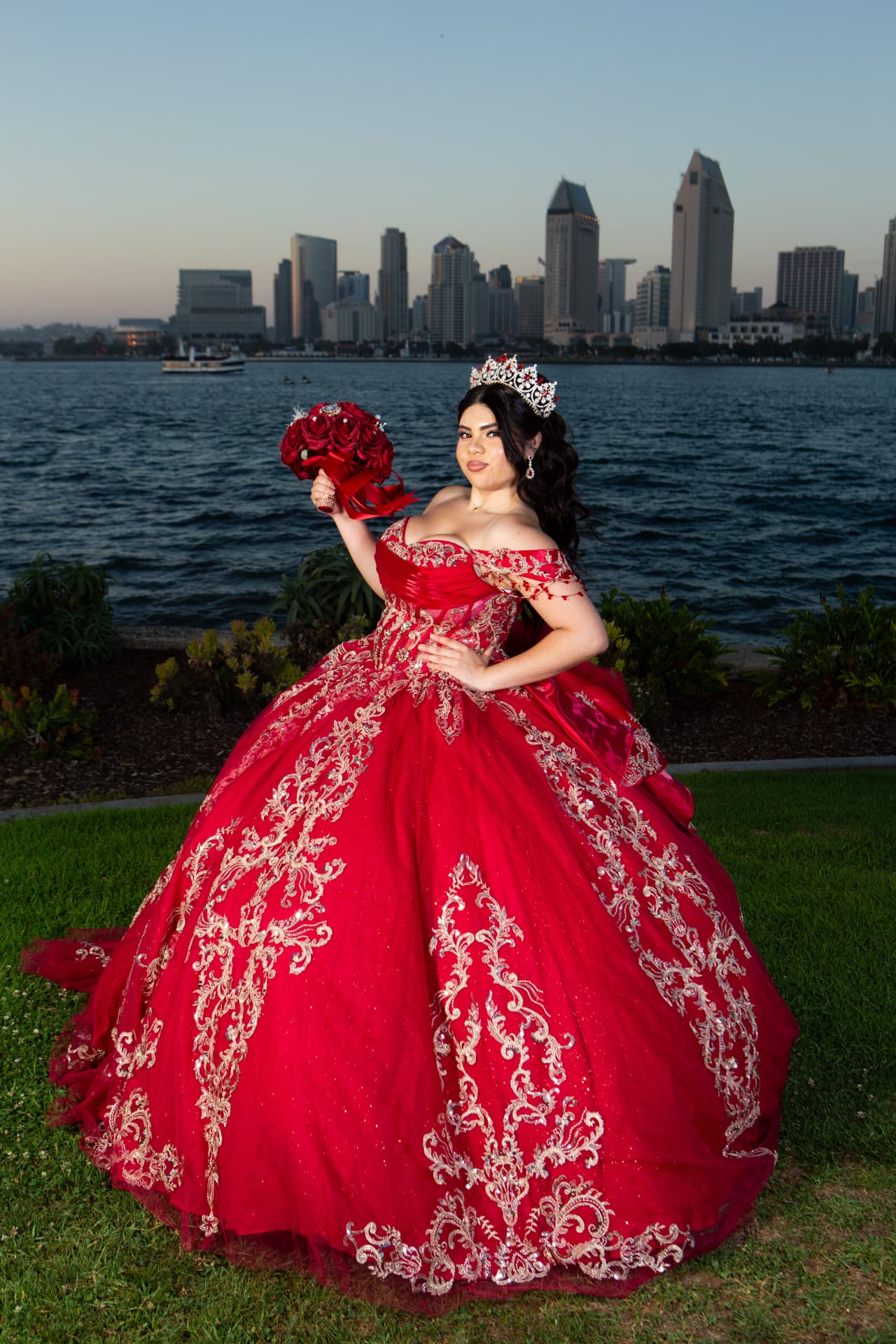 Quinceañera in a red gown holding her bouquet with the city skyline behind her at dusk