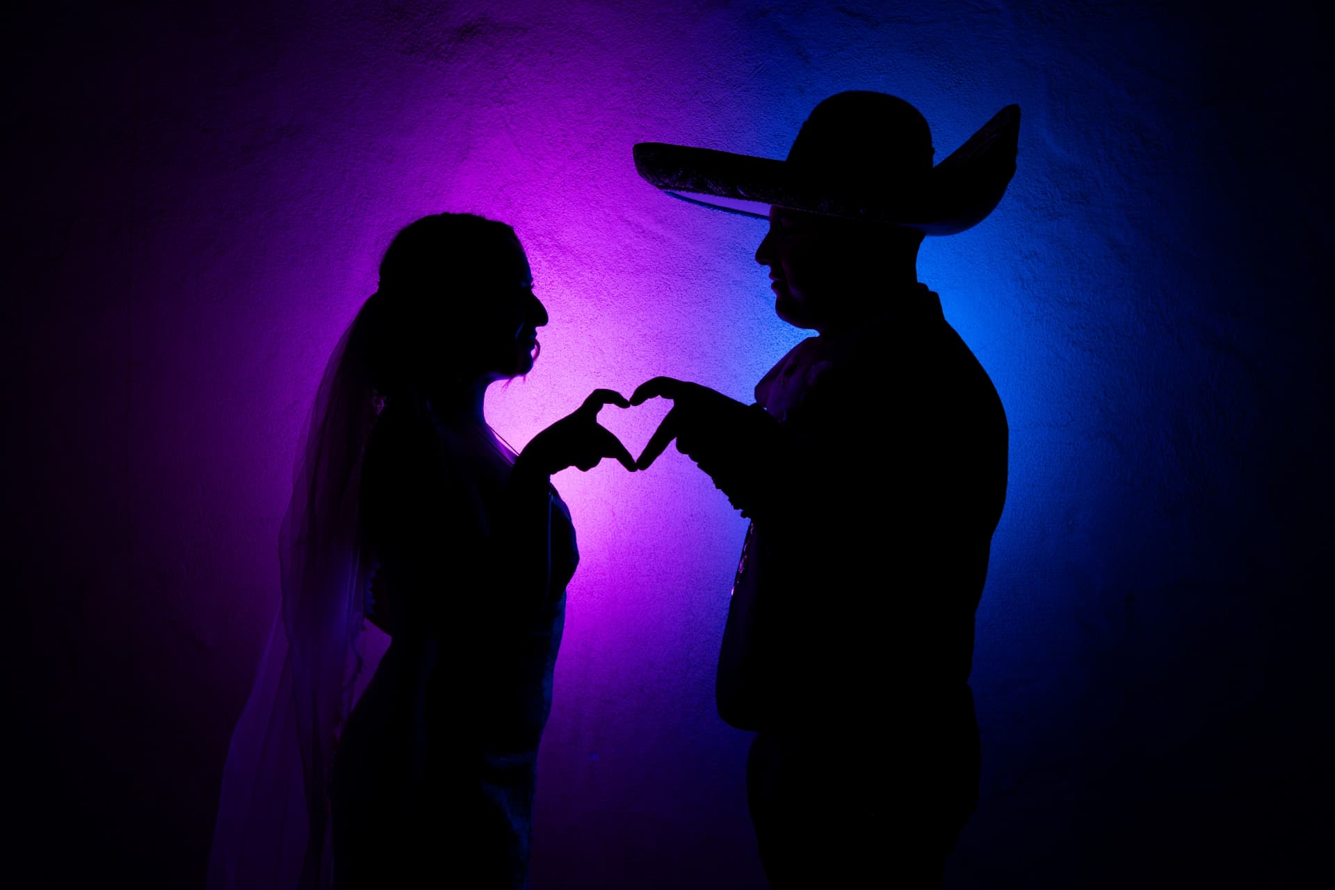 Silhouetted bride and groom forming a heart with their hands against magenta and blue light