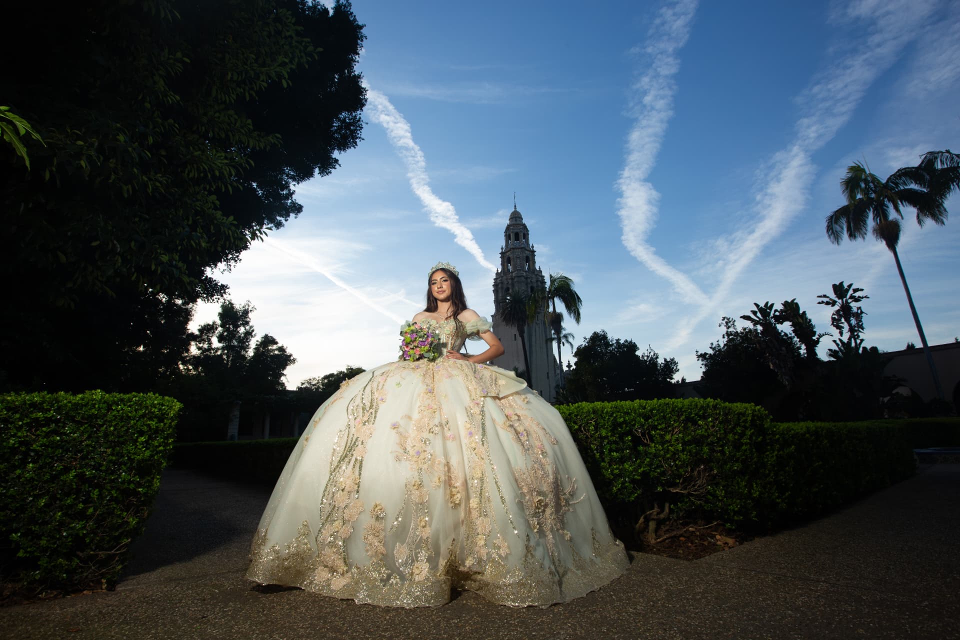 Quinceañera in an embroidered cream gown standing in a garden before the Balboa Park tower