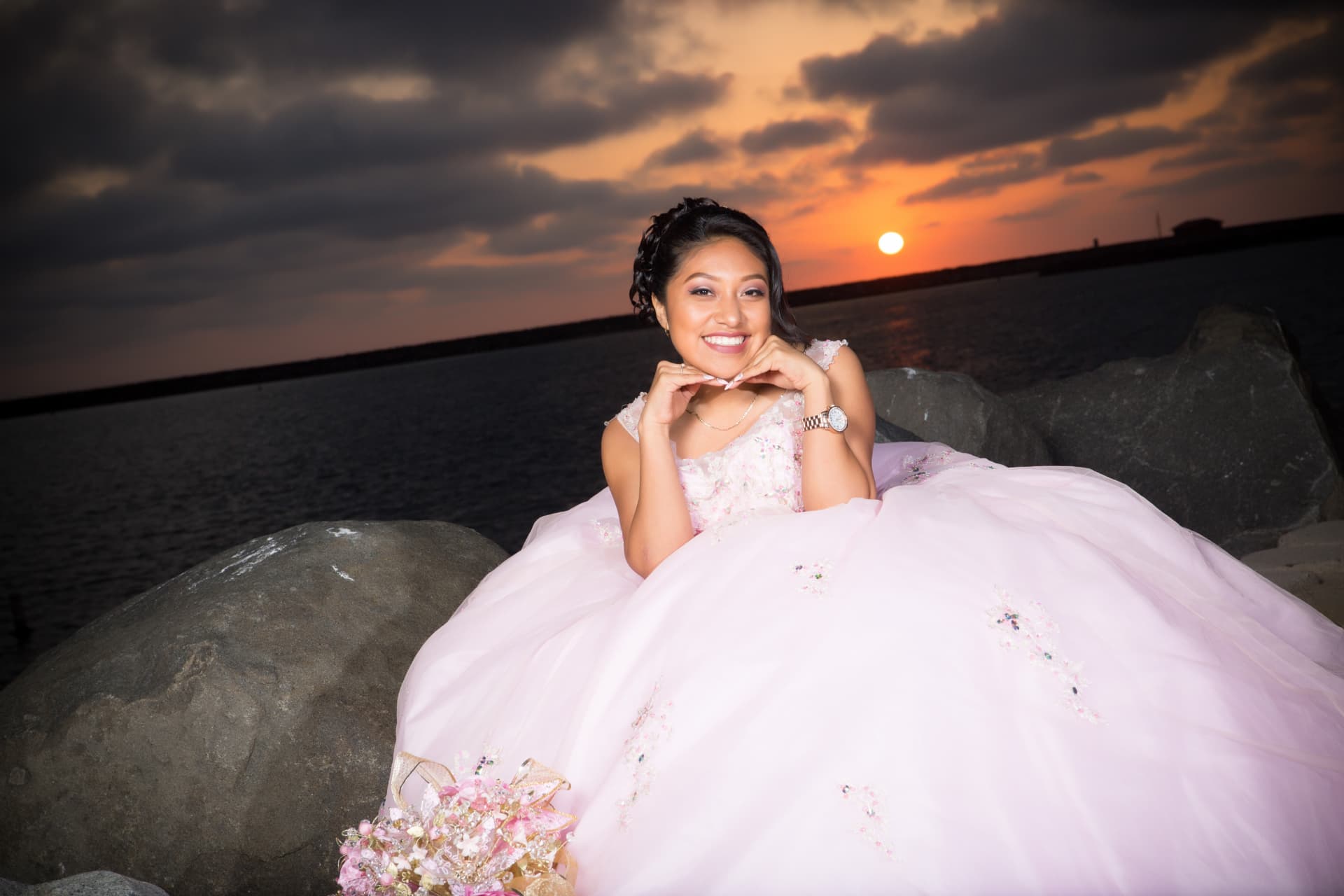 Quinceañera in a blush gown smiling beside breakwater rocks under a vivid sunset