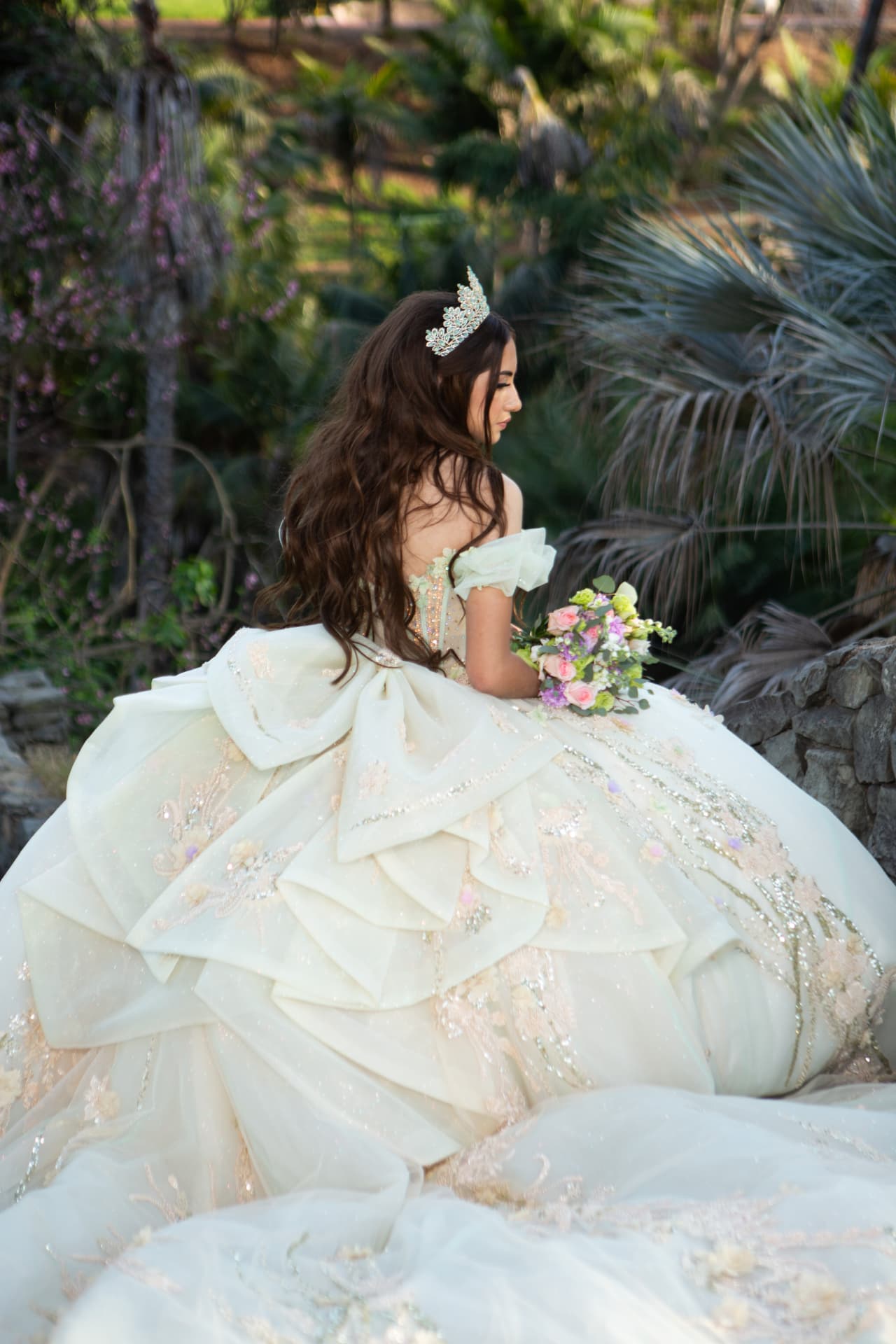 Quinceañera seen from behind in an ivory gown with a large layered bow and a bouquet